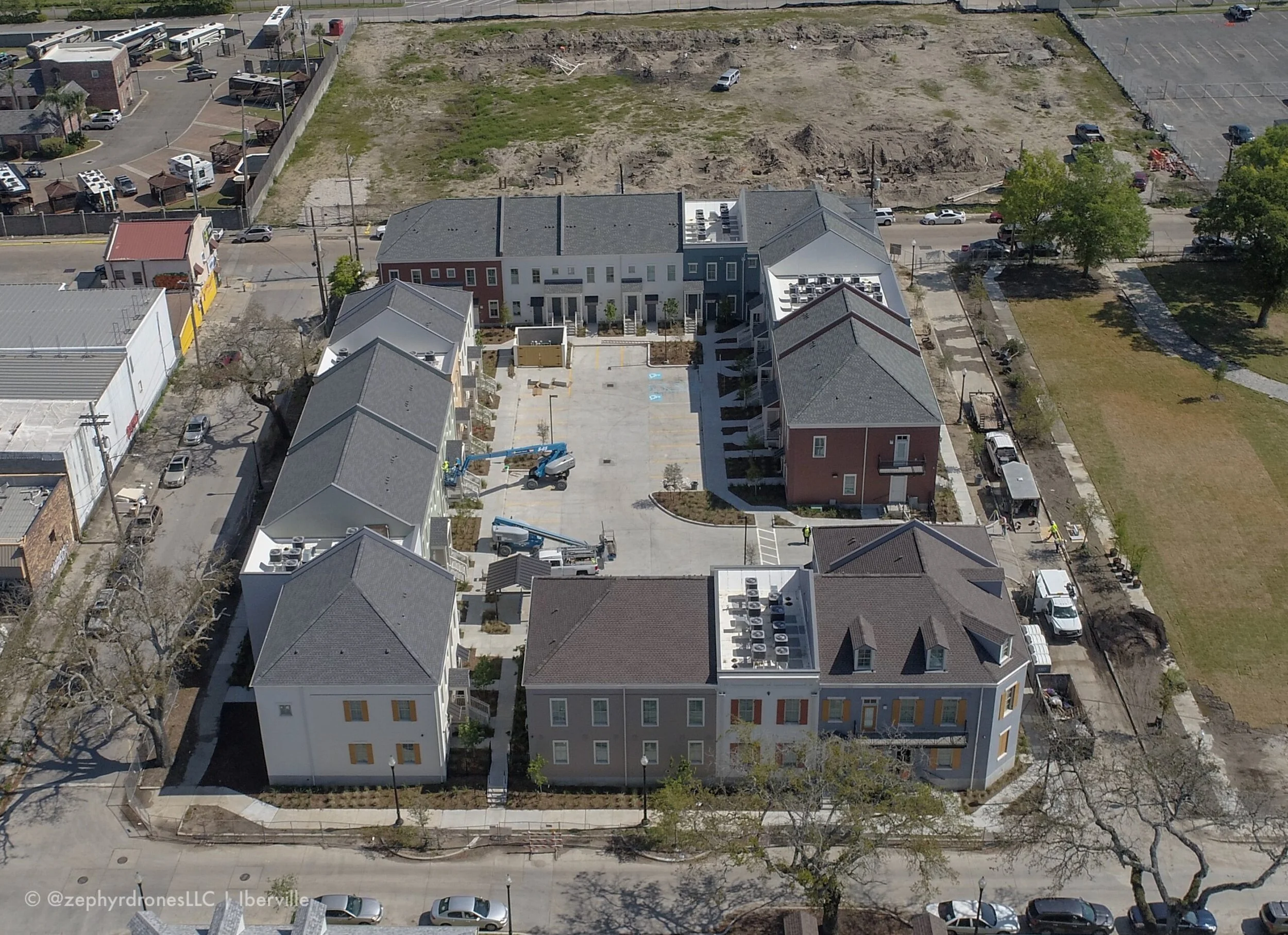 An aerial view of a newly constructed apartment complex with multiple buildings, parking lot, and construction equipment, with undeveloped land and some trees nearby.