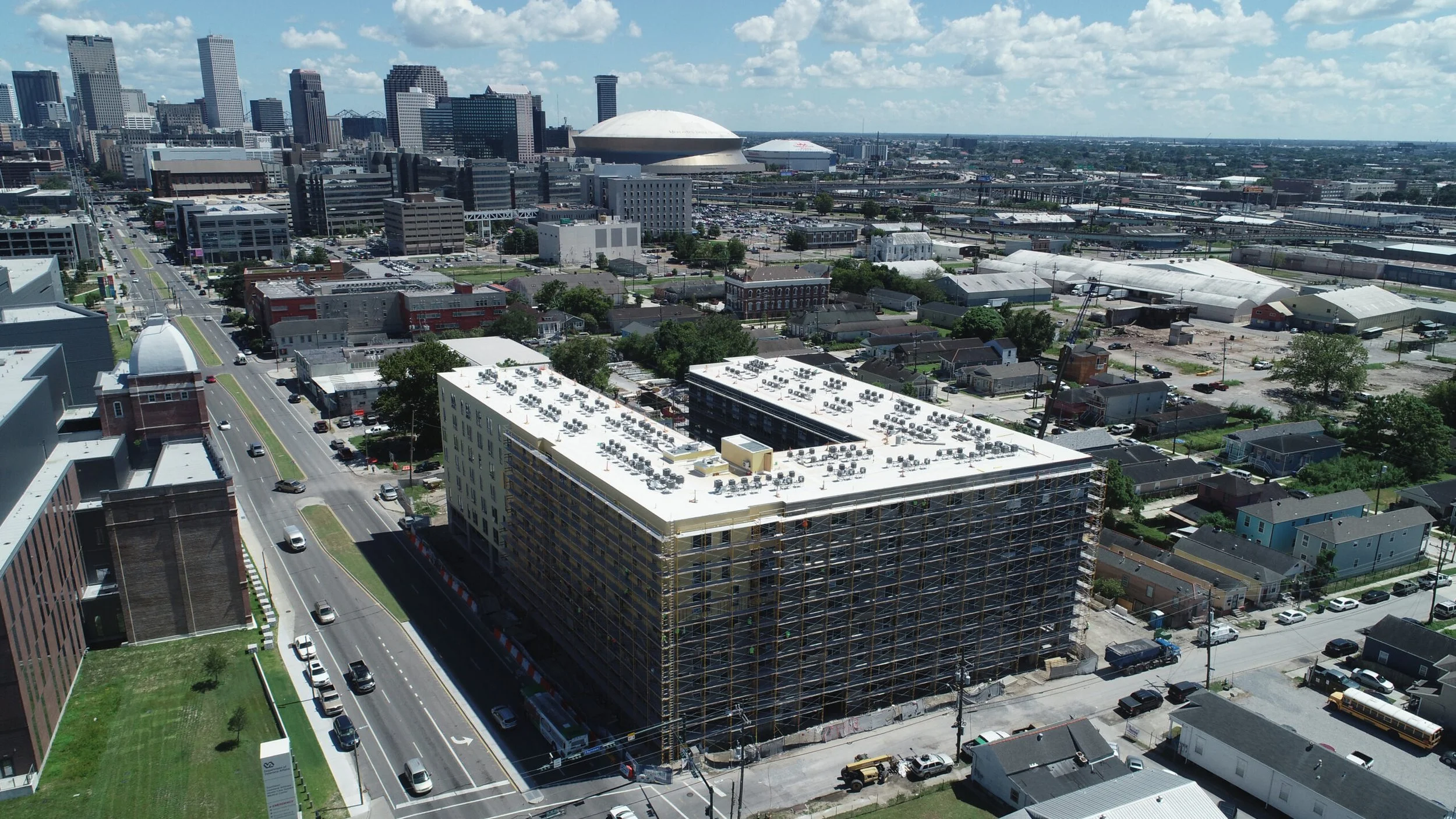 Aerial view of downtown cityscape with a large building under construction, featuring scaffolding, surrounded by streets, smaller buildings, and a skyline with high-rise buildings and a large domed stadium in the background.