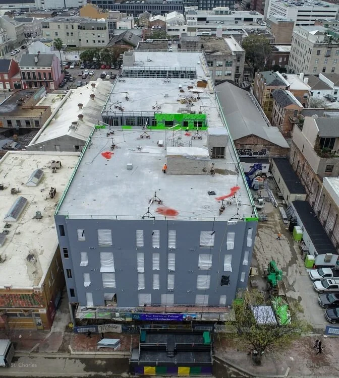 Aerial view of a multi-story building under construction in an urban area, with green scaffolding on the rooftop and construction workers visible. Surrounding buildings and parking lot are also visible.