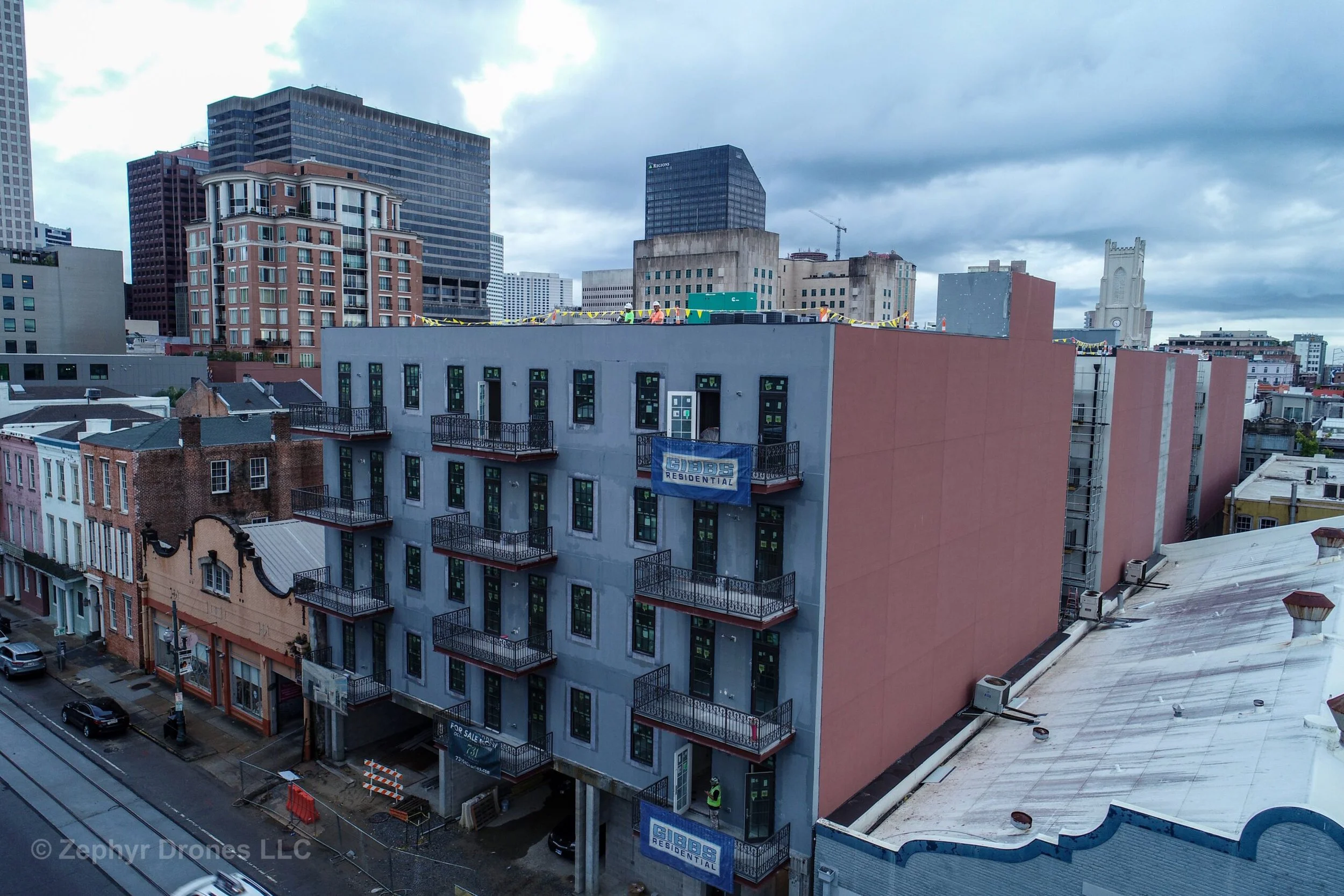 Urban cityscape with a multi-story building under construction, surrounded by taller buildings, under a cloudy sky.