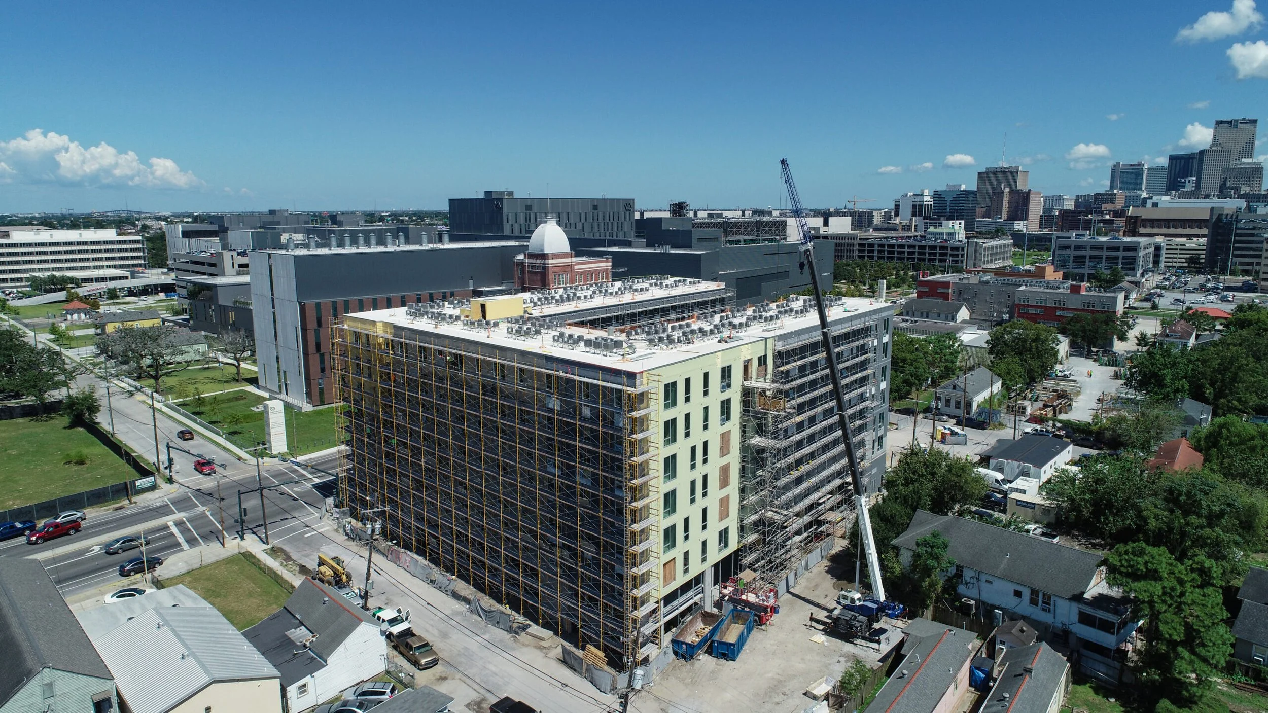 Aerial view of a construction site with a building under construction, scaffolding, a crane, and surrounding cityscape with taller buildings and blue sky.