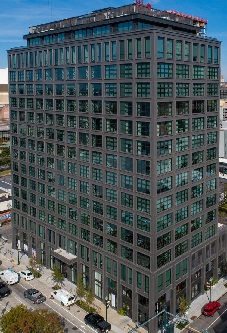 A tall modern office building with a dark gray facade and large windows, situated in an urban area with cars parked along the street and some trees.