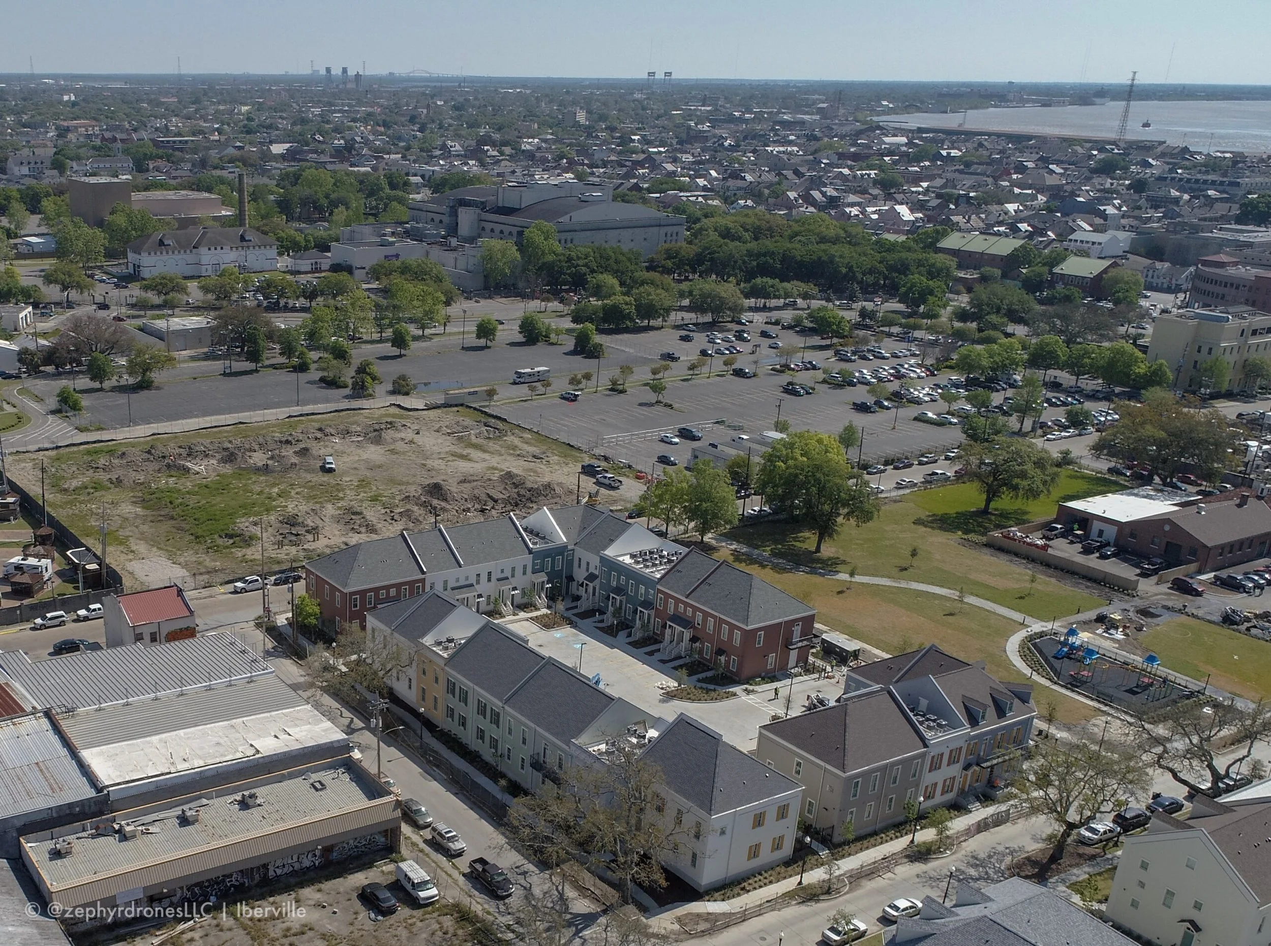 Aerial view of a cityscape with residential buildings, parking lots, green spaces, and a body of water in the background.
