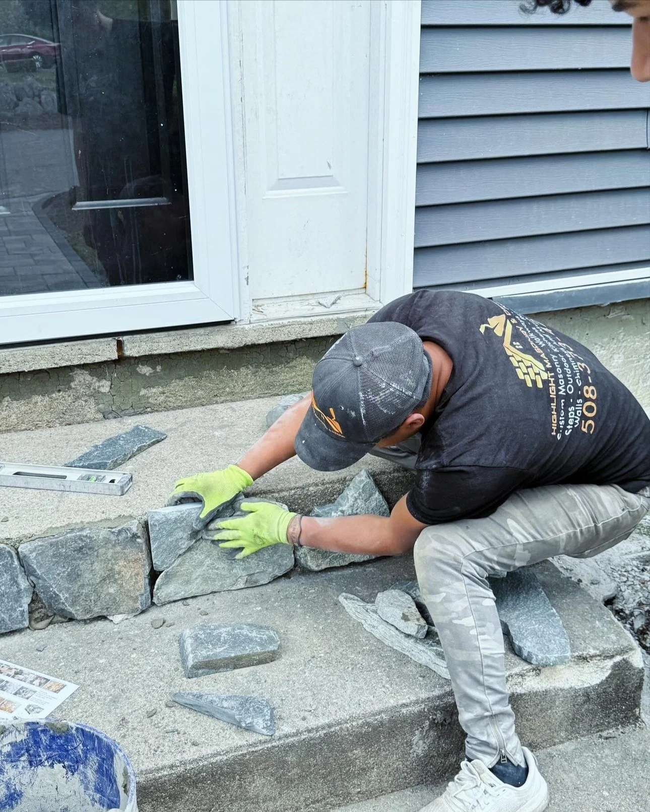 A person wearing a gray cap, black shirt, beige pants, and yellow gloves installing gray stone bricks on a concrete sidewalk outside a house with gray siding and a window.