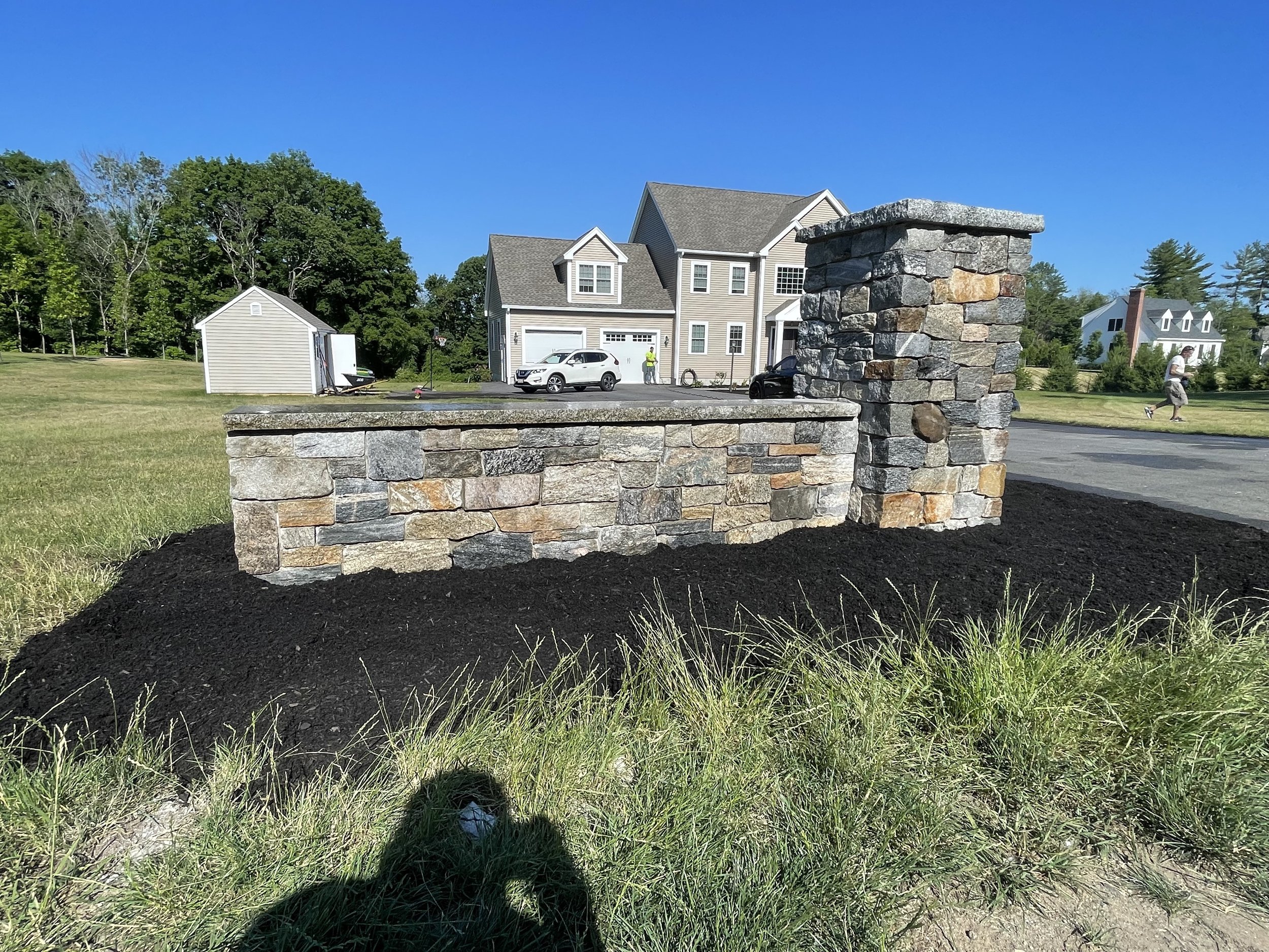 A decorative stone wall with pillars in a grassy yard, with houses and trees in the background on a sunny day.
