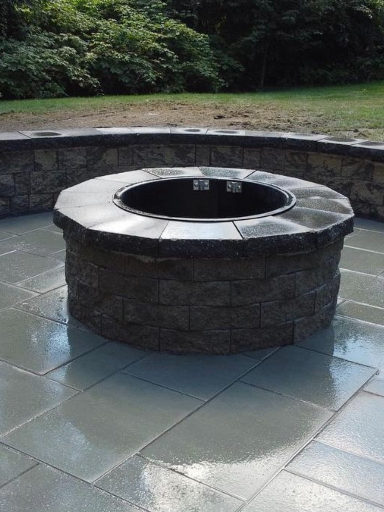 A circular fire pit with a black interior and a stone exterior, situated on a glistening tiled patio with a stone wall behind it and a grassy area with trees in the background.