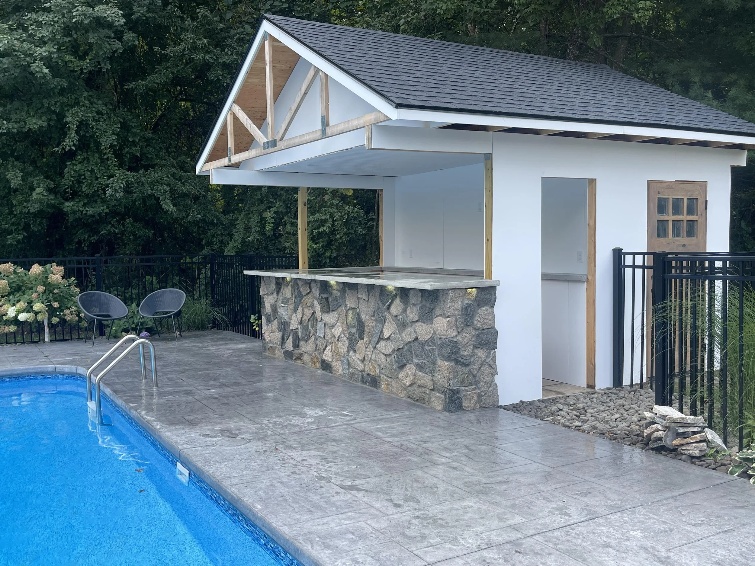Partially constructed poolside cabana with stone base, white walls, and wooden roof trusses, next to a blue swimming pool, surrounded by a black metal fence and greenery.