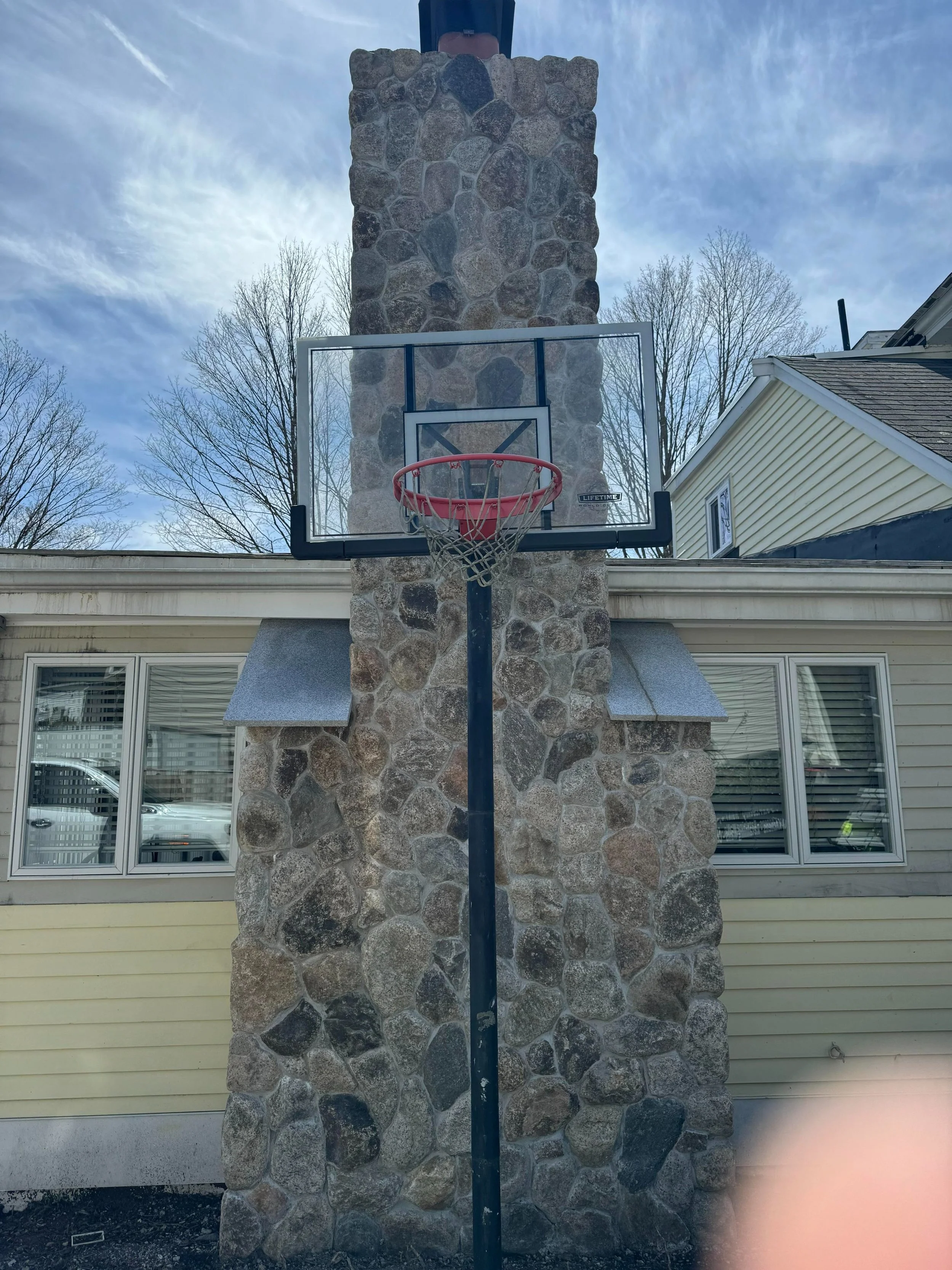 Outdoor basketball hoop mounted on a stone chimney with a house and blue sky in the background.