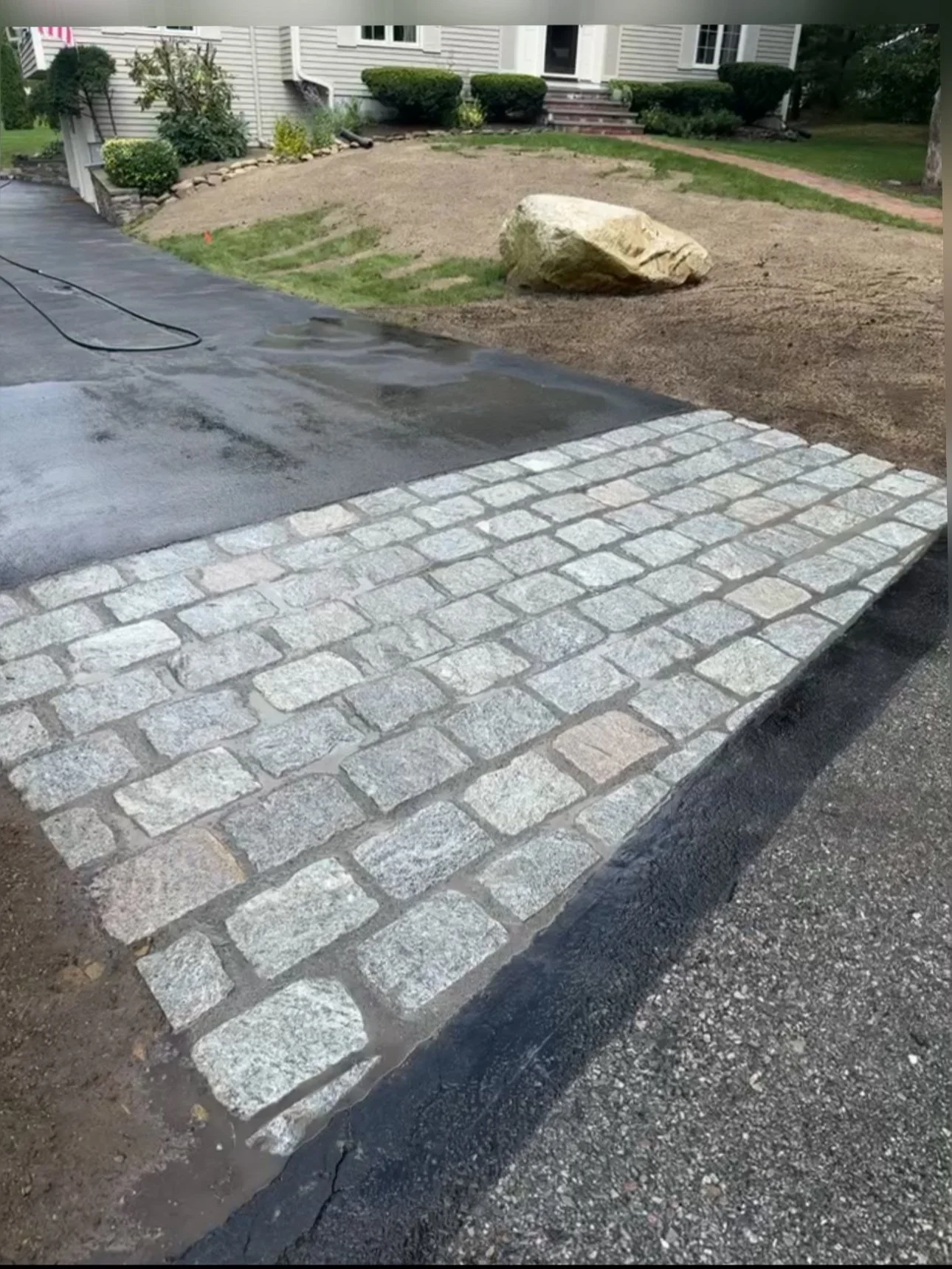 A newly paved driveway with cobblestone border leading to a house surrounded by a lawn and landscaping, with a large rock and patches of dirt visible in the yard.