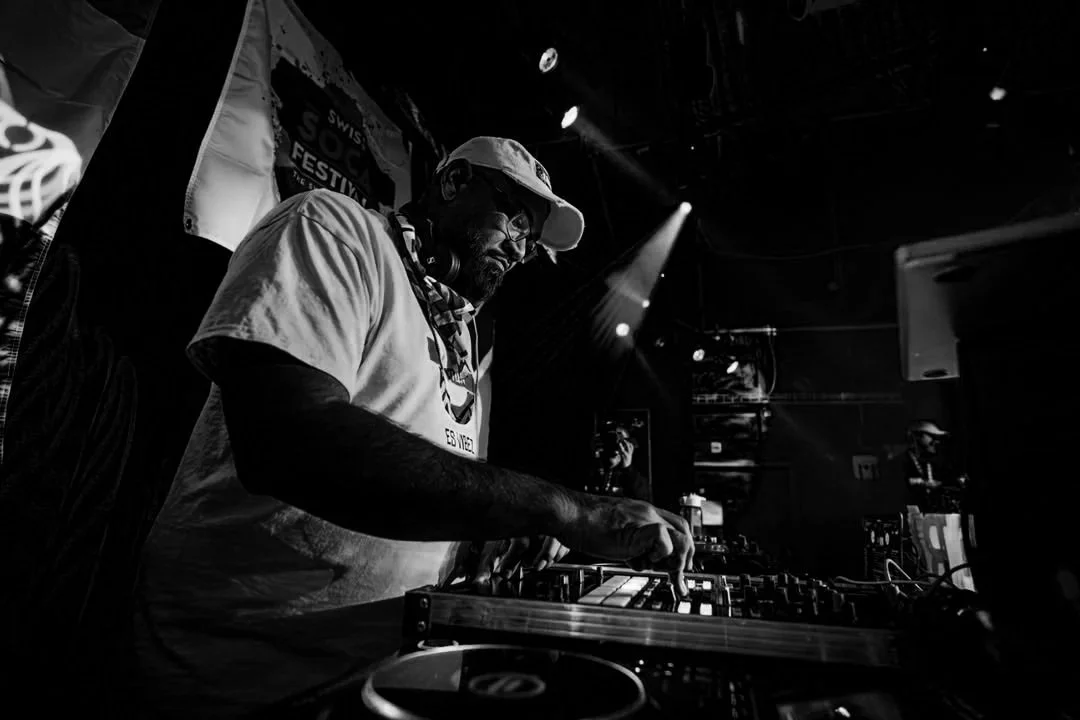 A DJ wearing a cap, glasses, and t-shirt, mixing music with professional DJ equipment on stage during a live event.