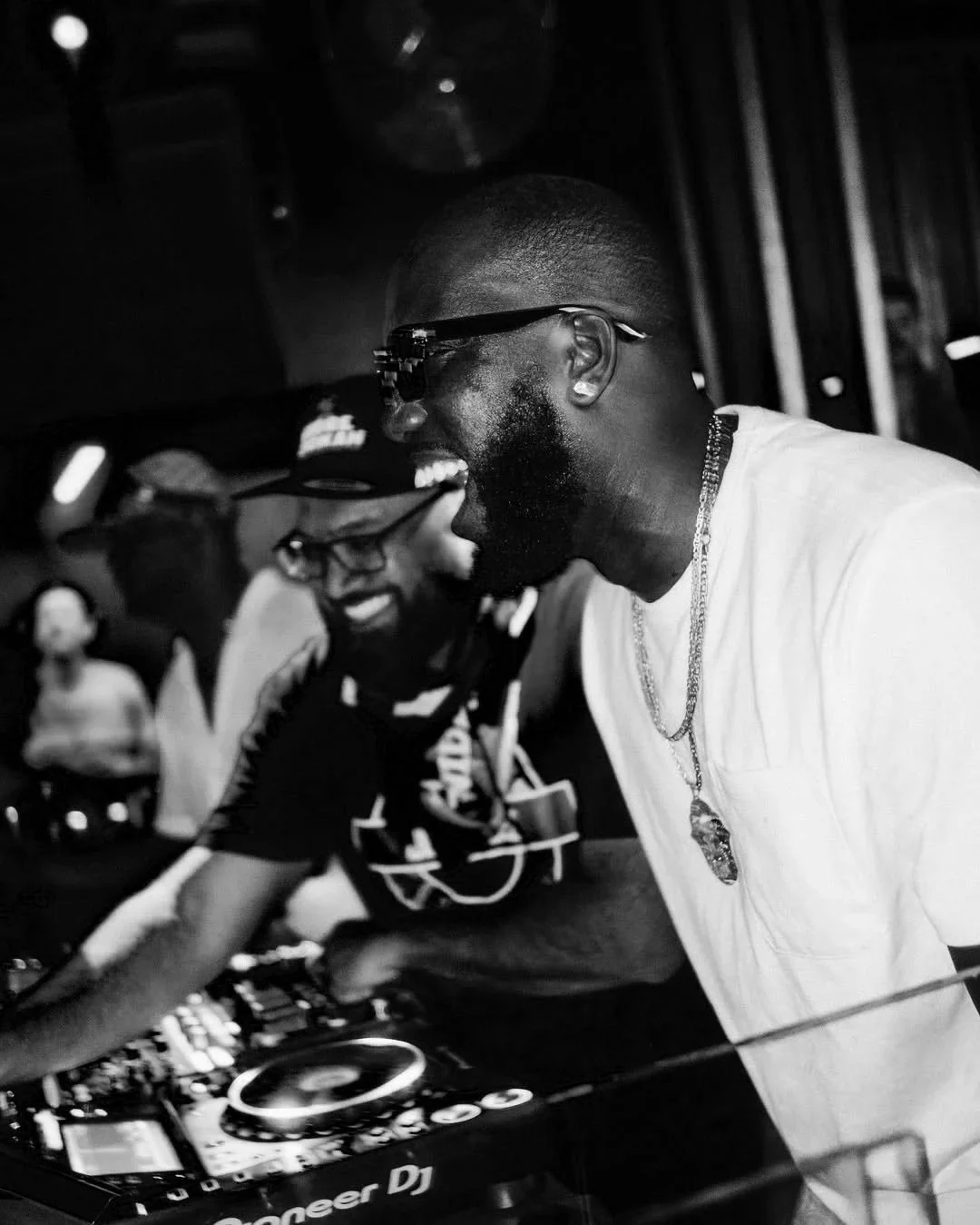 Two men are laughing and enjoying themselves at a DJ setup in a club or party setting, with one man wearing glasses, a necklace, and a white shirt, and the other in a hat, glasses, and a patterned shirt.