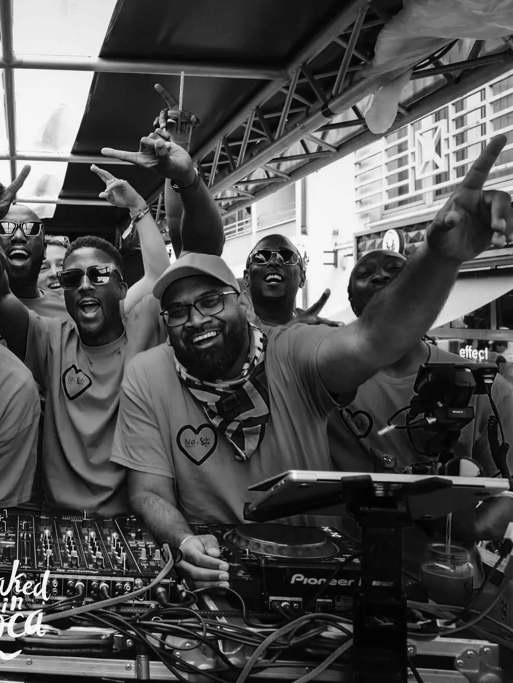 A group of people at a DJ booth, smiling and celebrating with some making peace and pointing gestures. The scene appears to be at an outdoor event or party, with a structure and city buildings in the background.