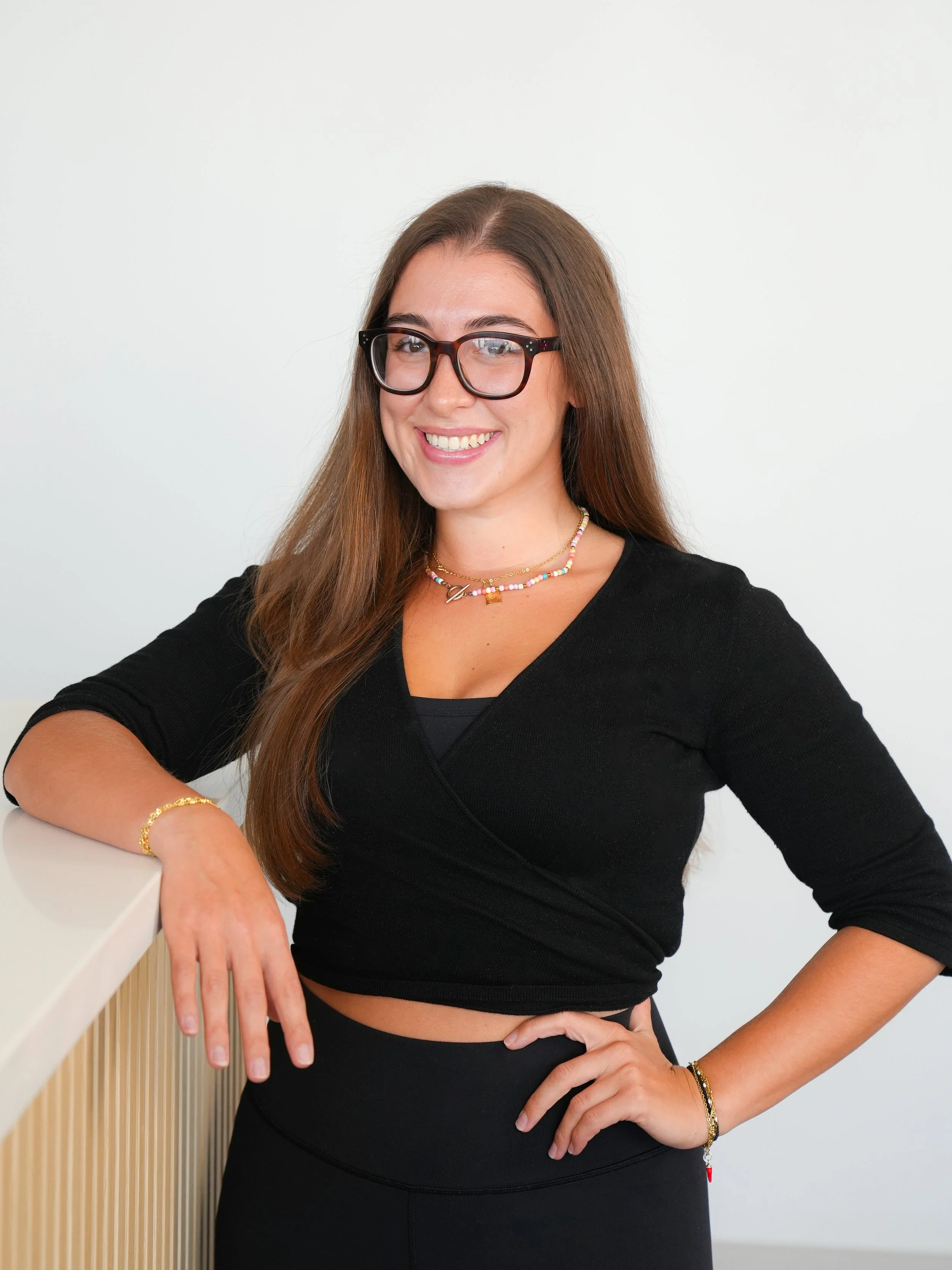 A woman with long brown hair wearing black glasses, a black wrap top, and layered colorful necklaces, standing with her left hand on her hip and her right arm resting on a white railing, smiling at the camera against a plain white wall.