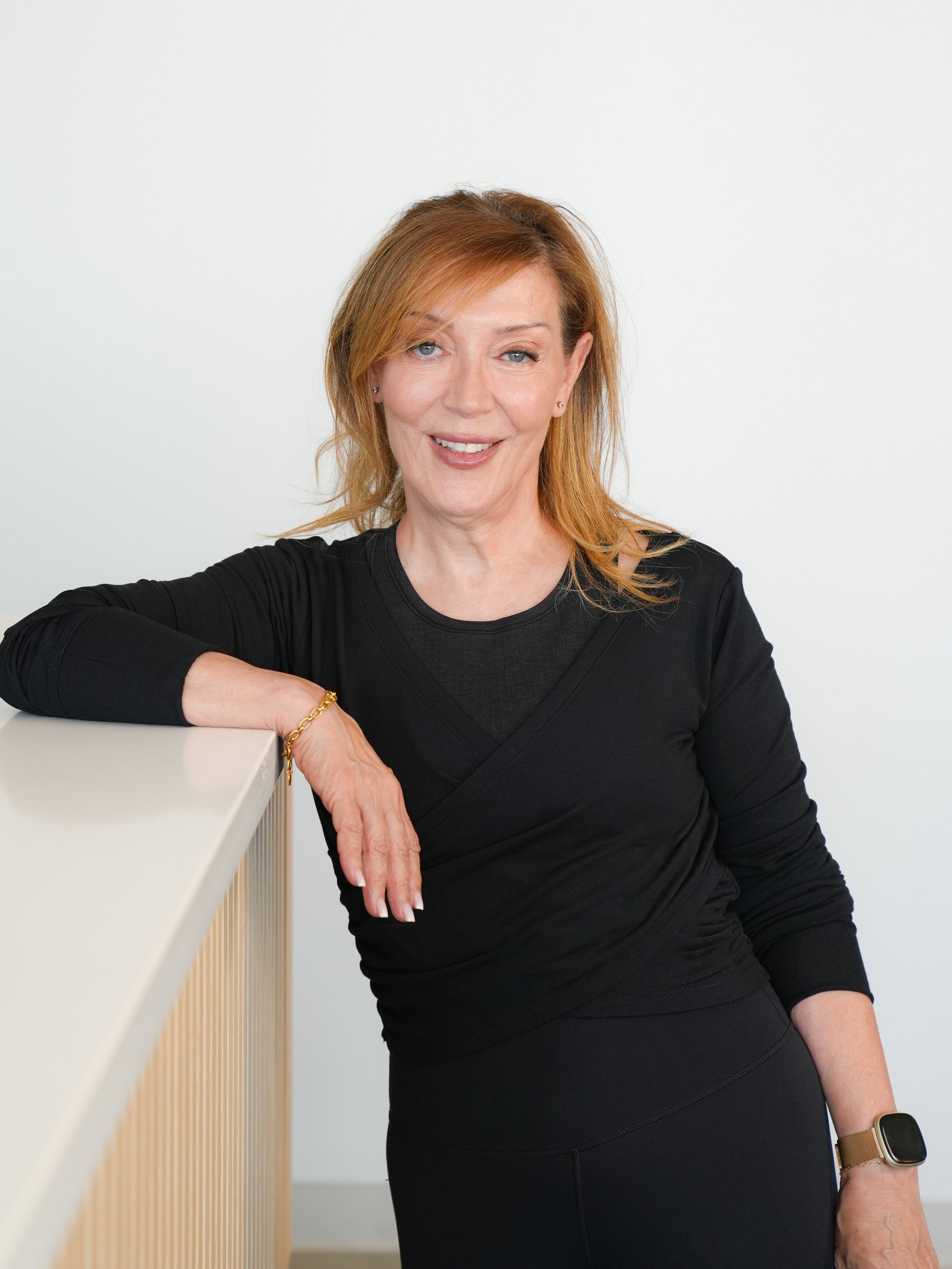 A woman with reddish hair wearing a black long-sleeve top and black skirt, leaning on a white surface with a wooden railing, looking at the camera with a slight smile, against a plain white background.