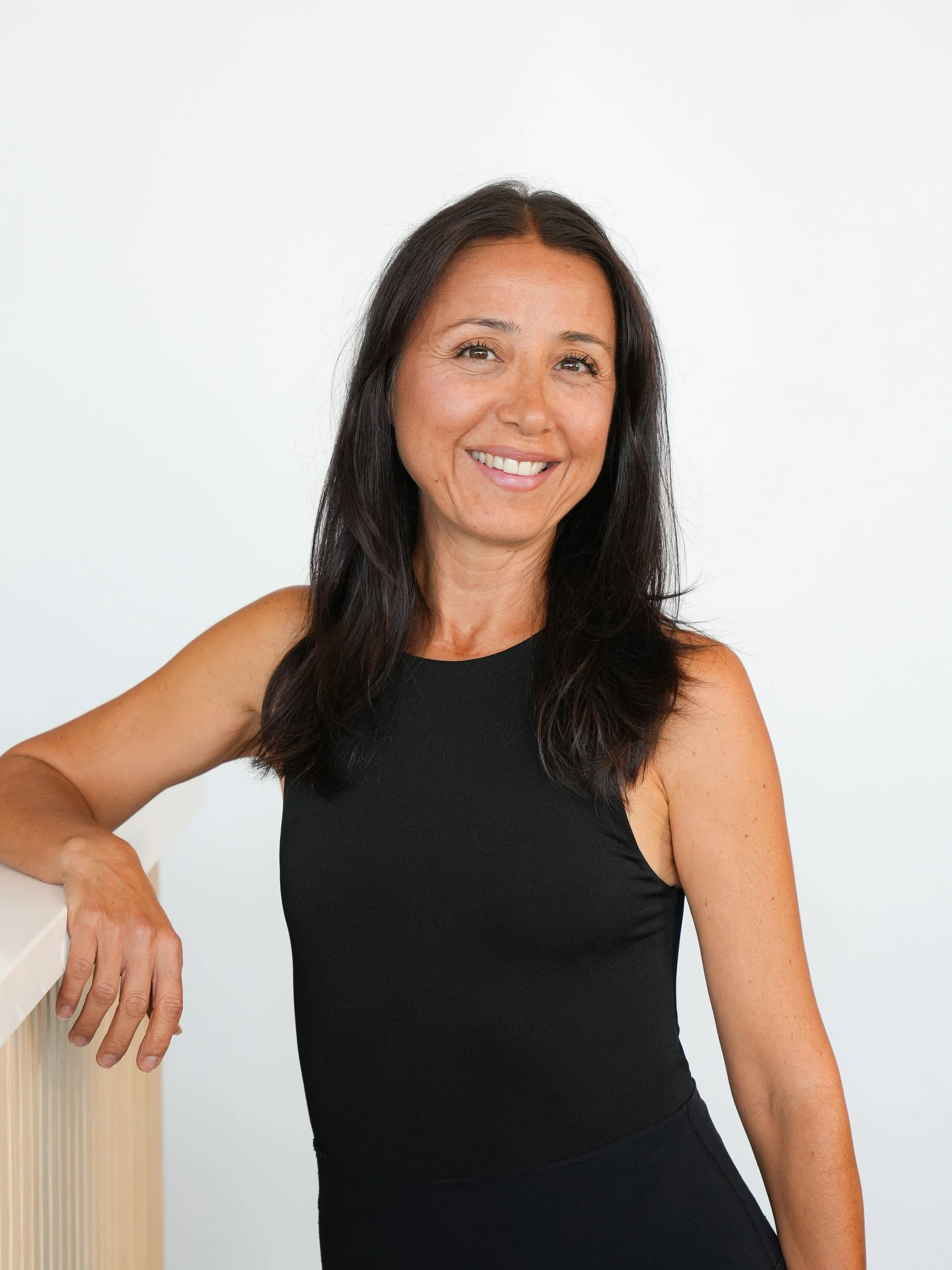 A woman with long dark hair smiling, wearing a black sleeveless top, leaning on a white railing with a plain white background.