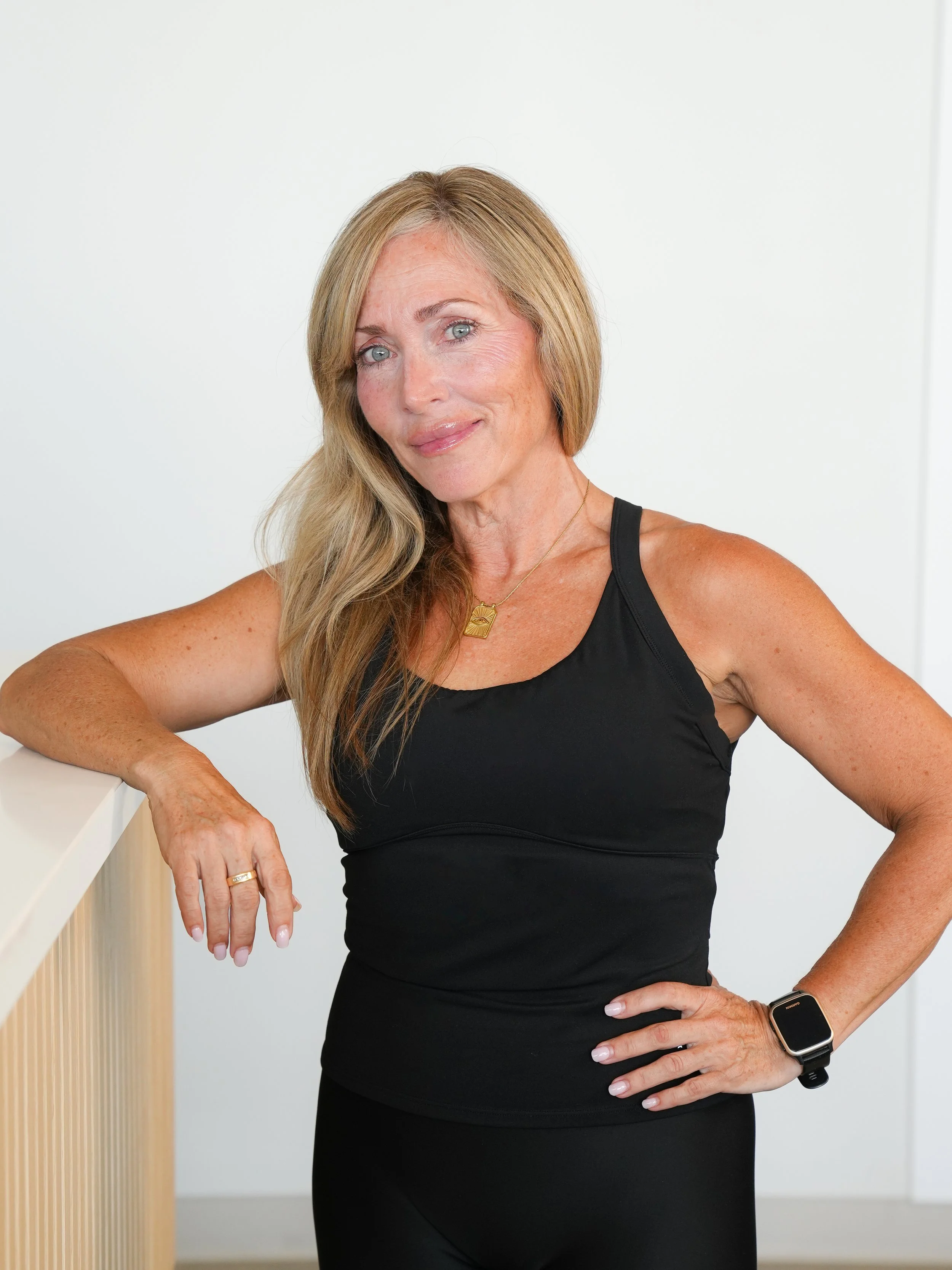 A woman with blonde hair wearing a black sleeveless top and a smartwatch standing indoors against a plain white wall, resting her left arm on a wooden surface and looking at the camera.