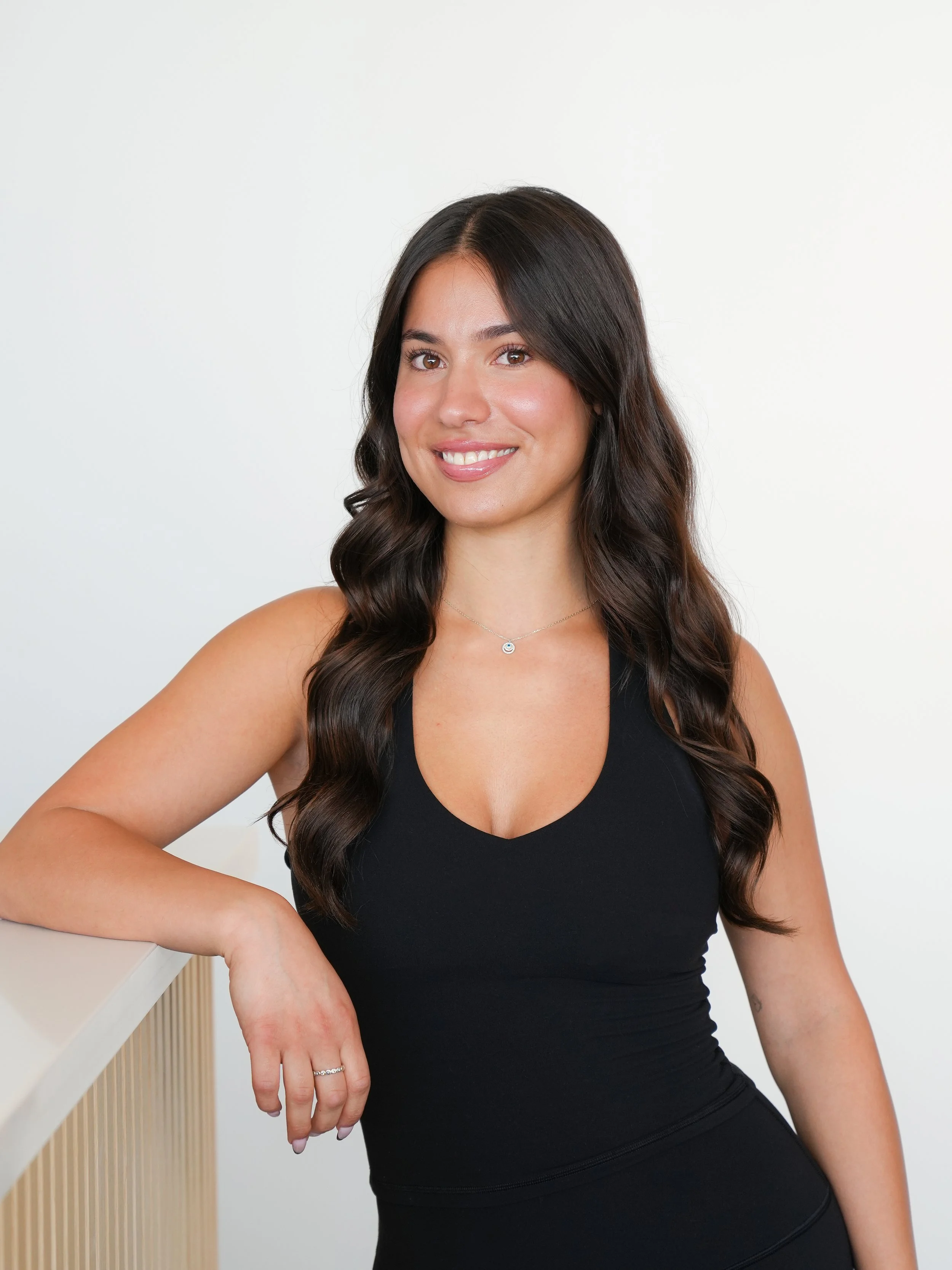 A woman with long dark hair in loose waves, wearing a black sleeveless top, smiling, and posing with her right arm resting on a white surface.