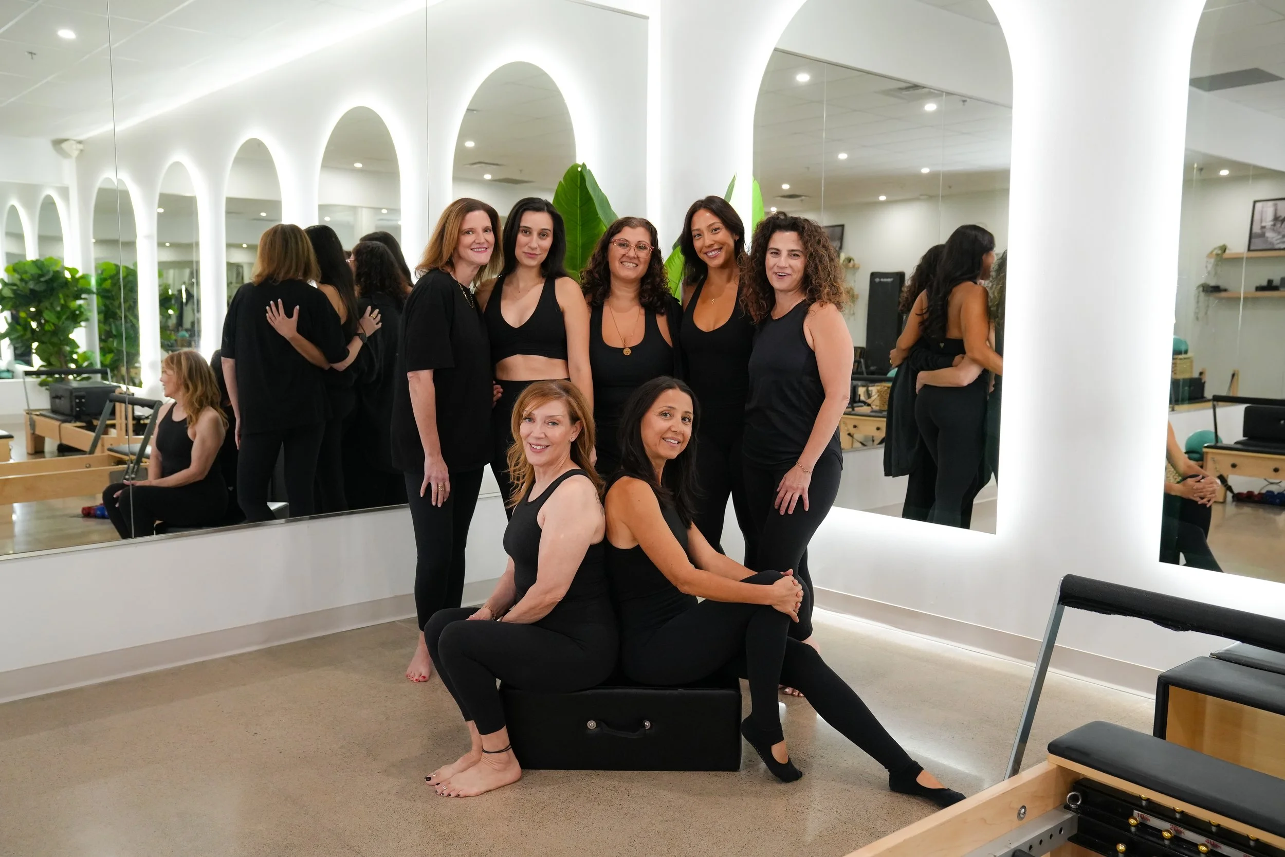 Group of seven women in black clothing gathered in a wellness or fitness studio with mirrored walls and indoor plants. Some are sitting, some are standing, smiling for the photo.