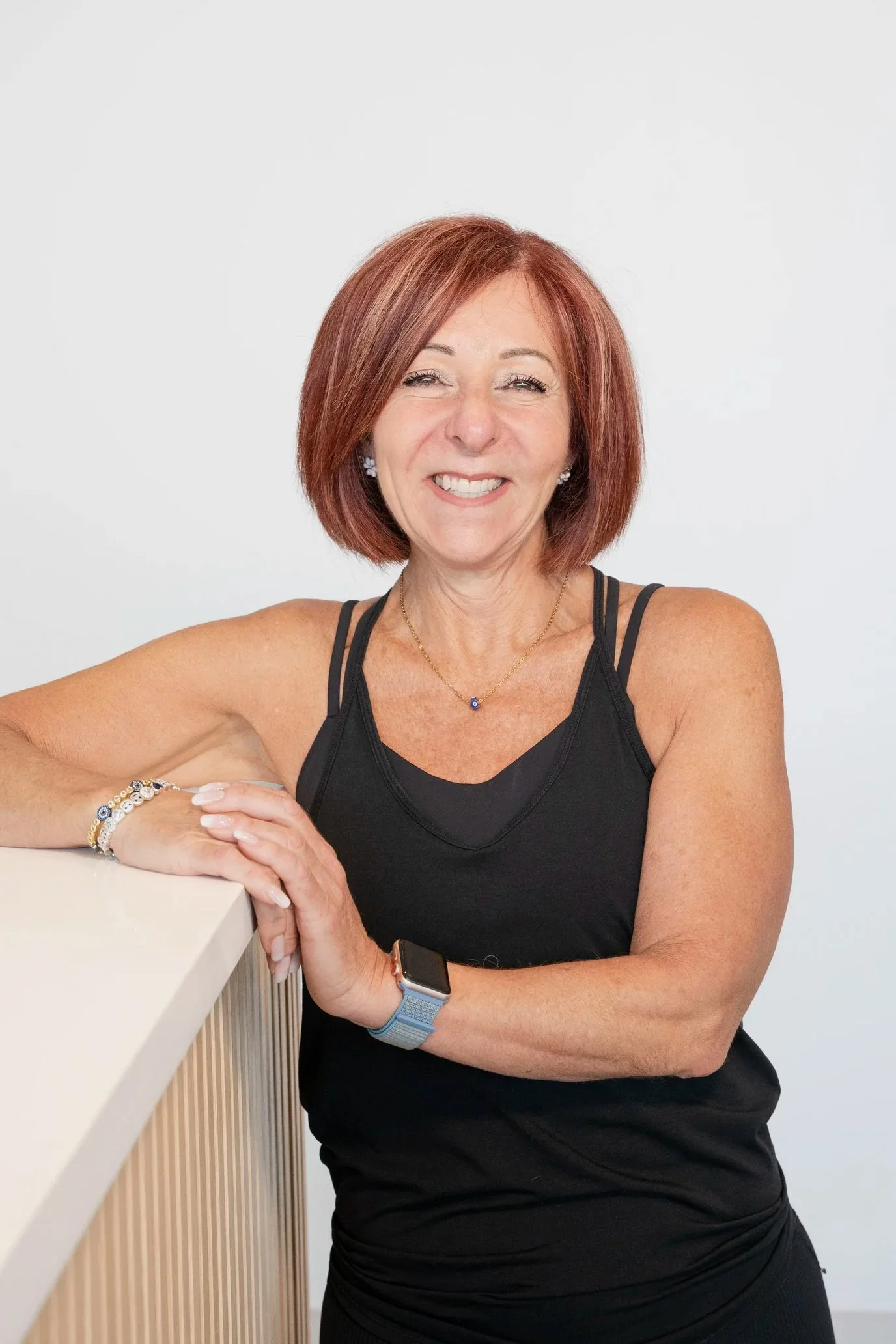 A smiling woman with red hair, wearing a black athletic top, jewelry, and a smartwatch, leaning on a white table against a plain white background.