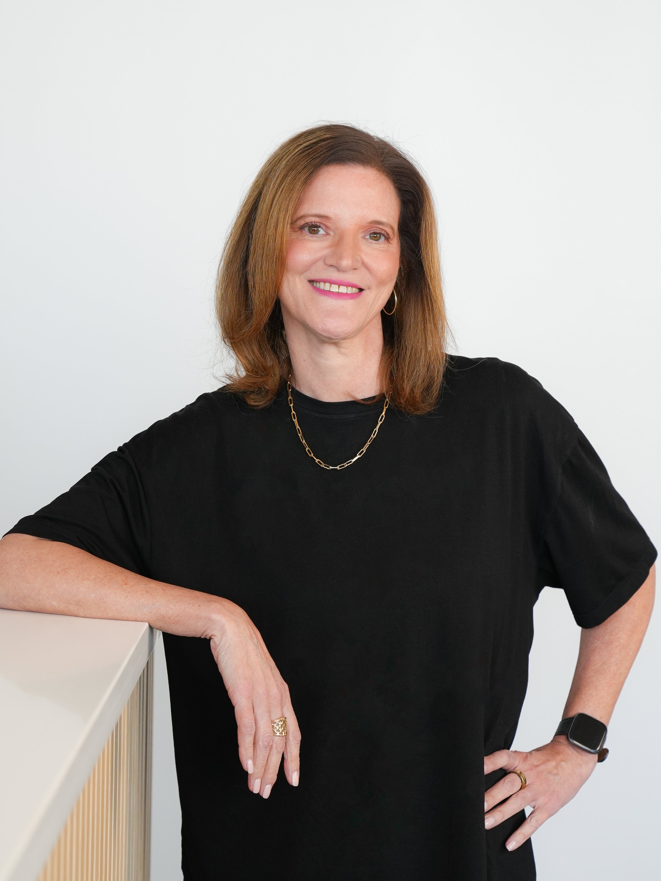 A woman with shoulder-length brown hair wearing a black shirt, gold necklace, hoop earrings, and an Apple smartwatch, leaning on a white surface against a white background, smiling at the camera.