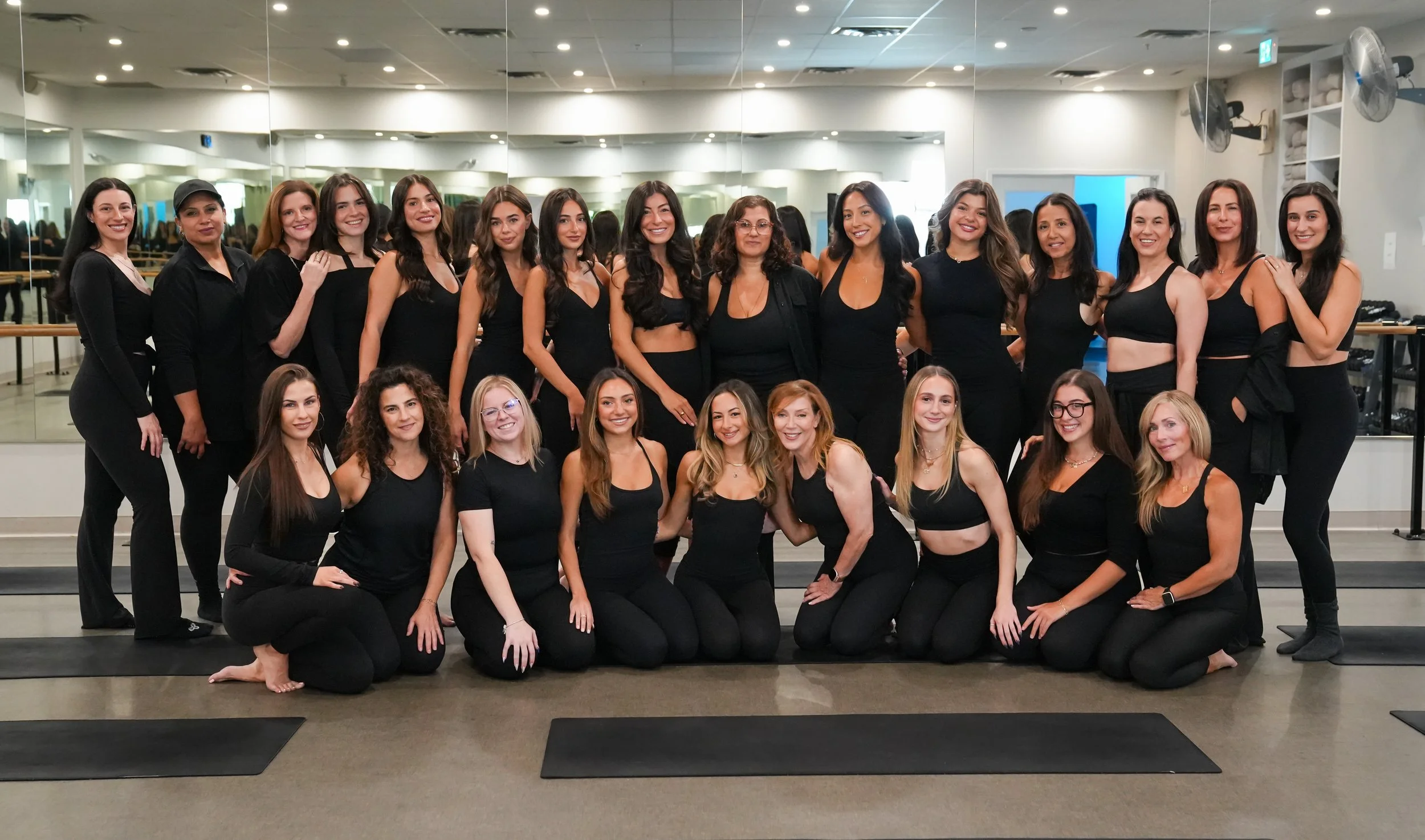 Group of women in black workout attire posing in a fitness studio gym with mirrors and ballet bars.