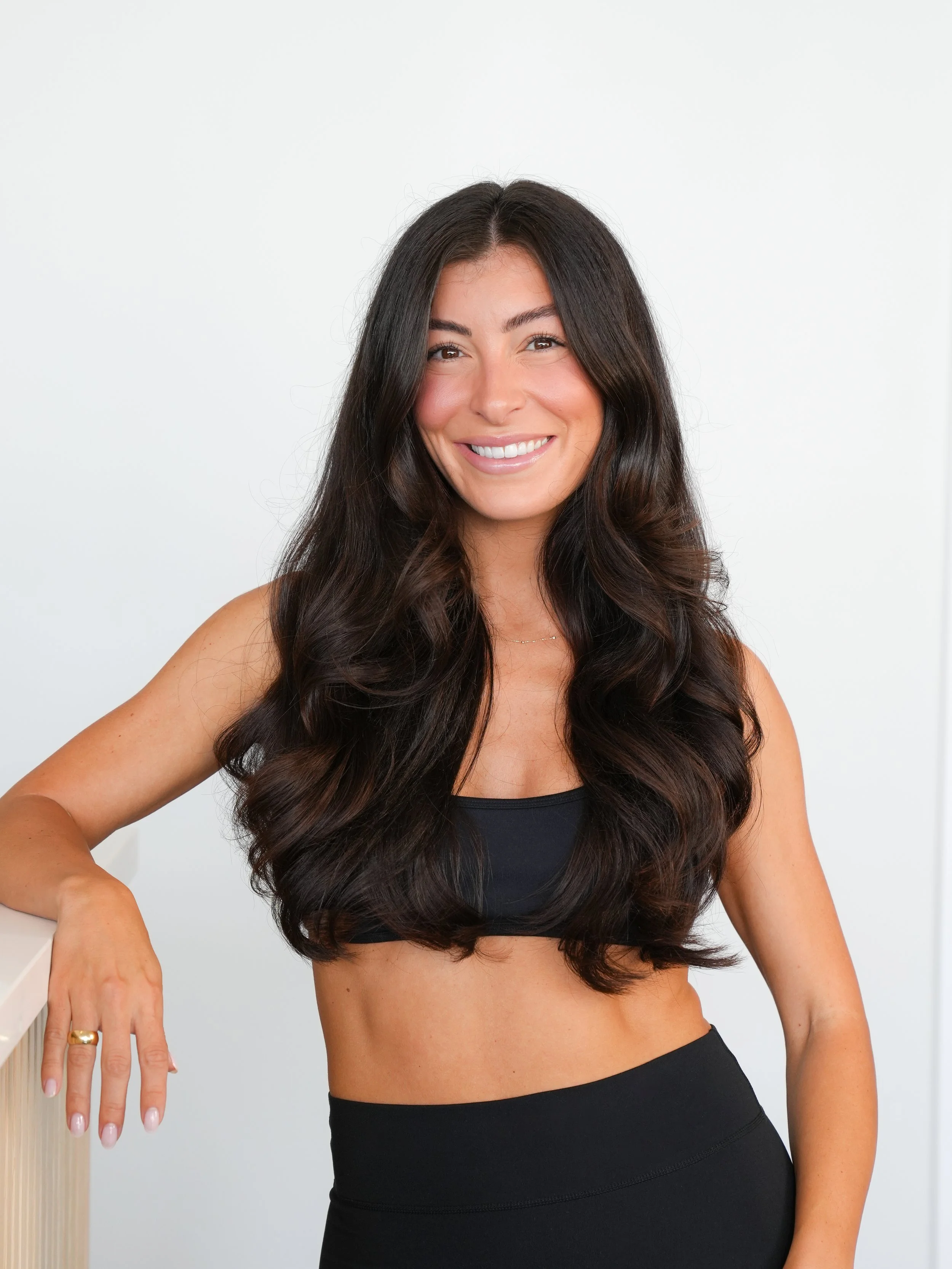 A woman with long, dark, wavy hair smiling and wearing a black sports bra and high-waisted shorts, standing against a plain white background.
