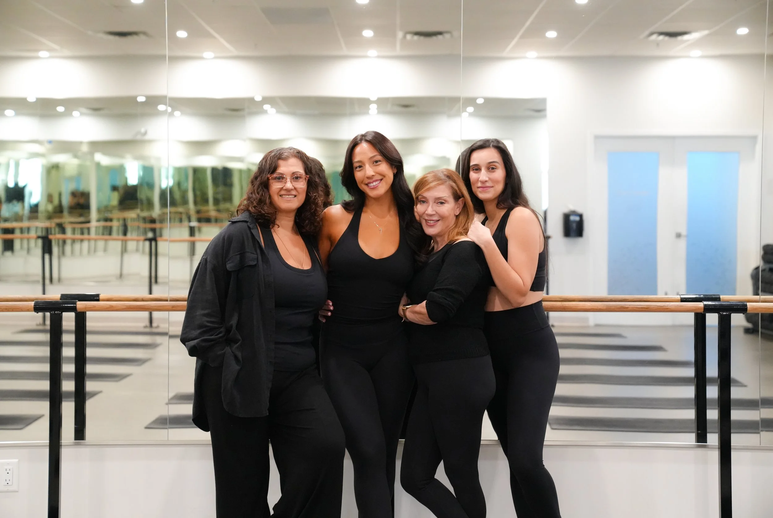 Group of four women smiling and posing in a dance studio in front of a mirrored wall with ballet barres.