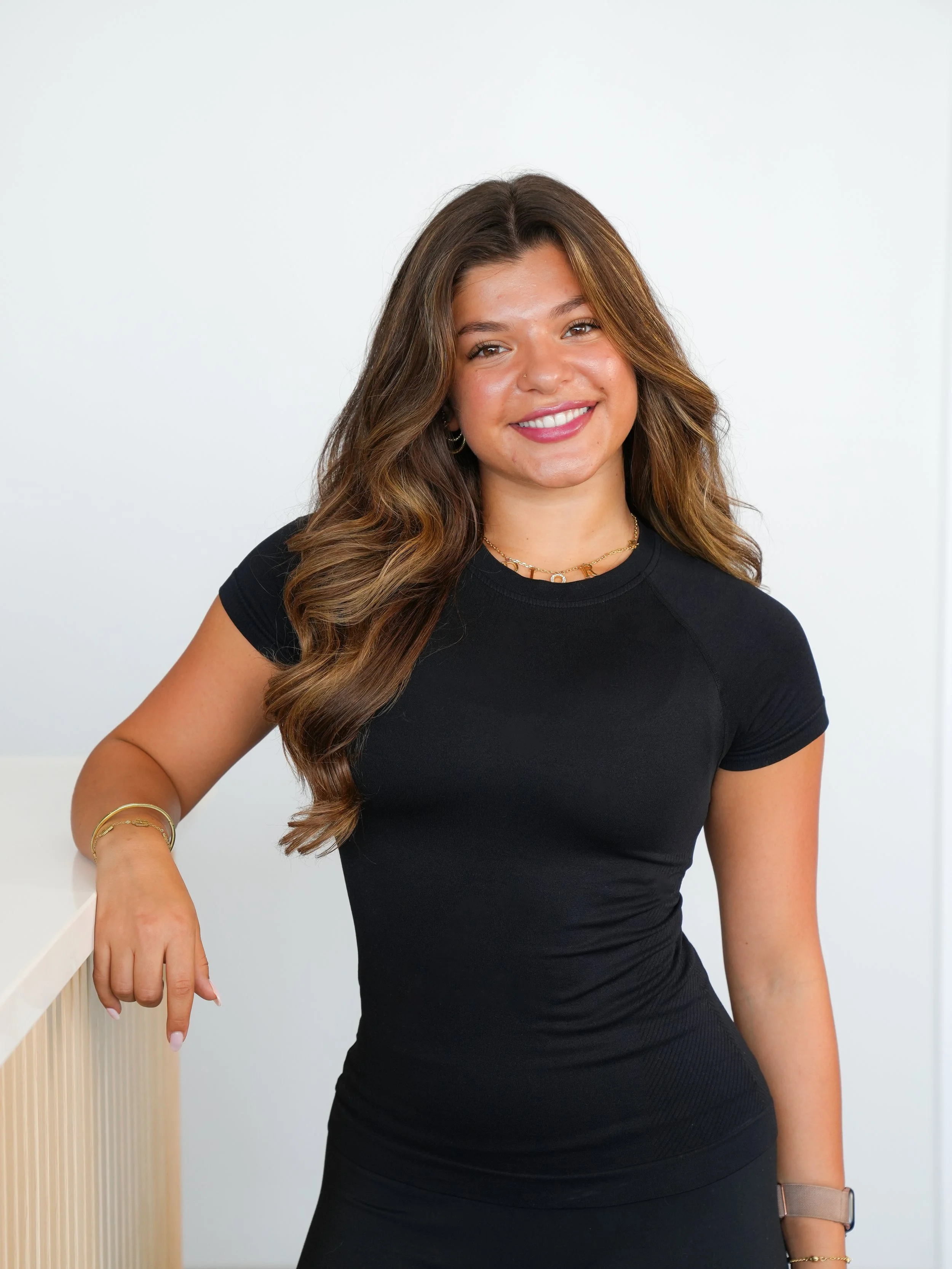 A smiling young woman with long wavy brown hair wearing a black fitted t-shirt, gold jewelry, and a smartwatch, standing indoors against a white wall.