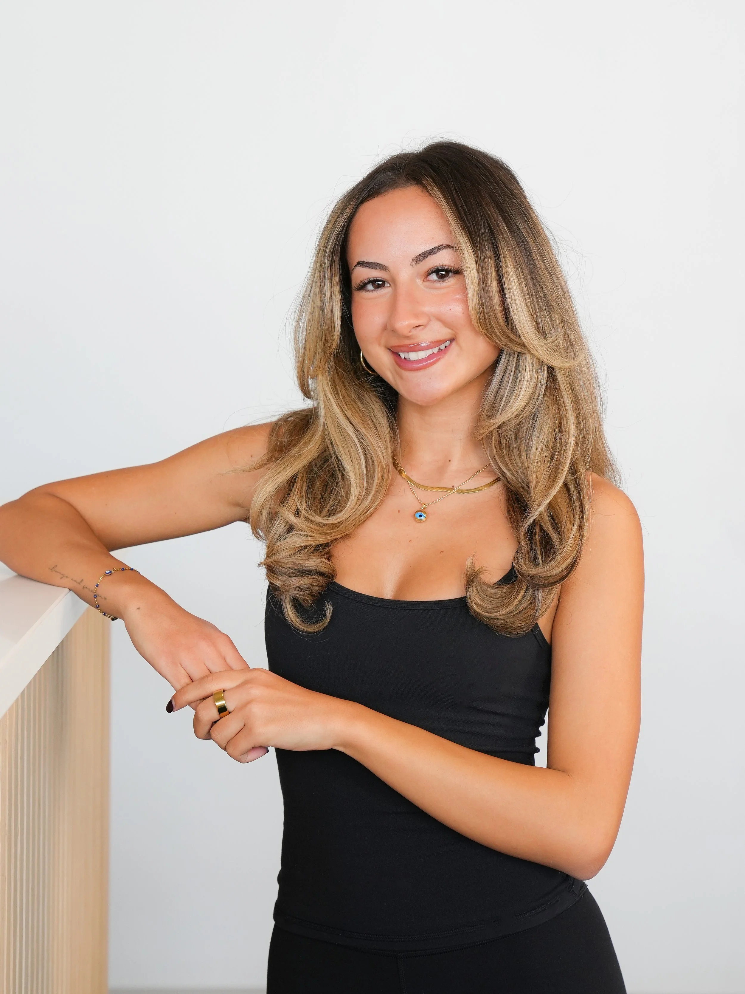 A young woman with long, wavy hair wearing a black sleeveless top and gold jewelry, smiling while leaning on a railing against a plain white background.