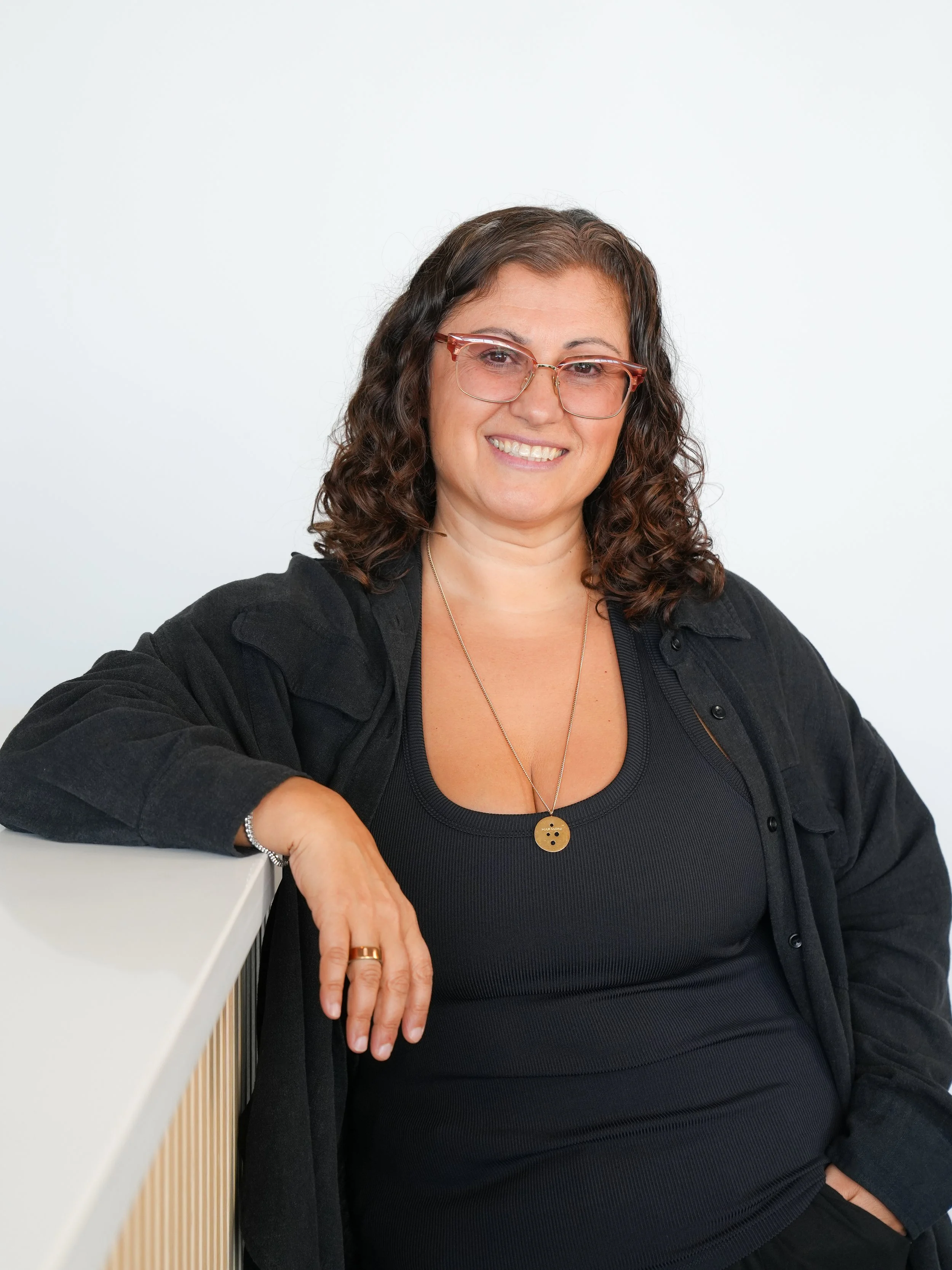 A woman with curly dark hair and glasses smiling, sitting at a white table against a plain light background, wearing a black top and jacket with a necklace and a bracelet.