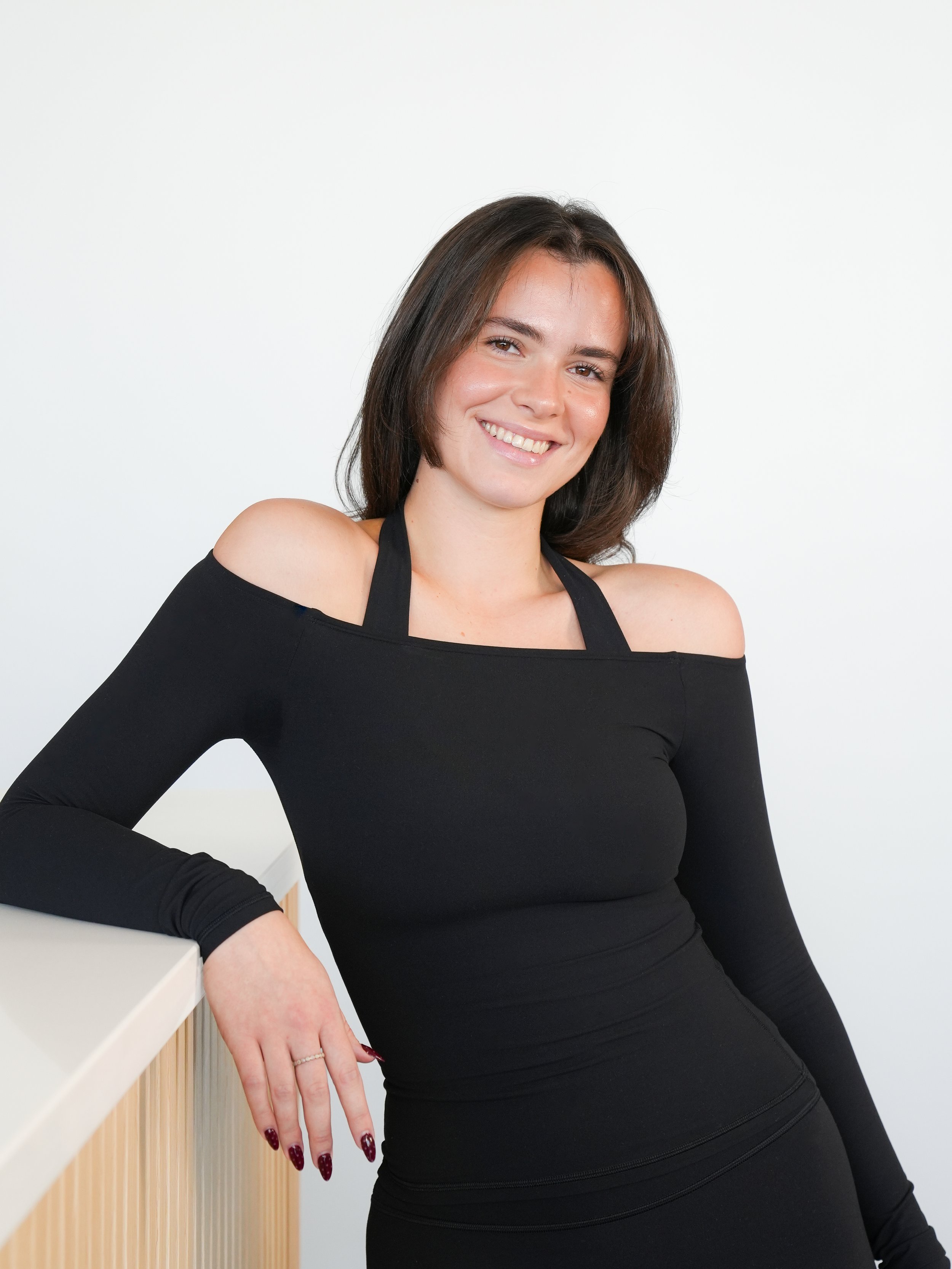 A woman with shoulder-length dark hair smiling, wearing a black off-shoulder top, standing against a plain white background.