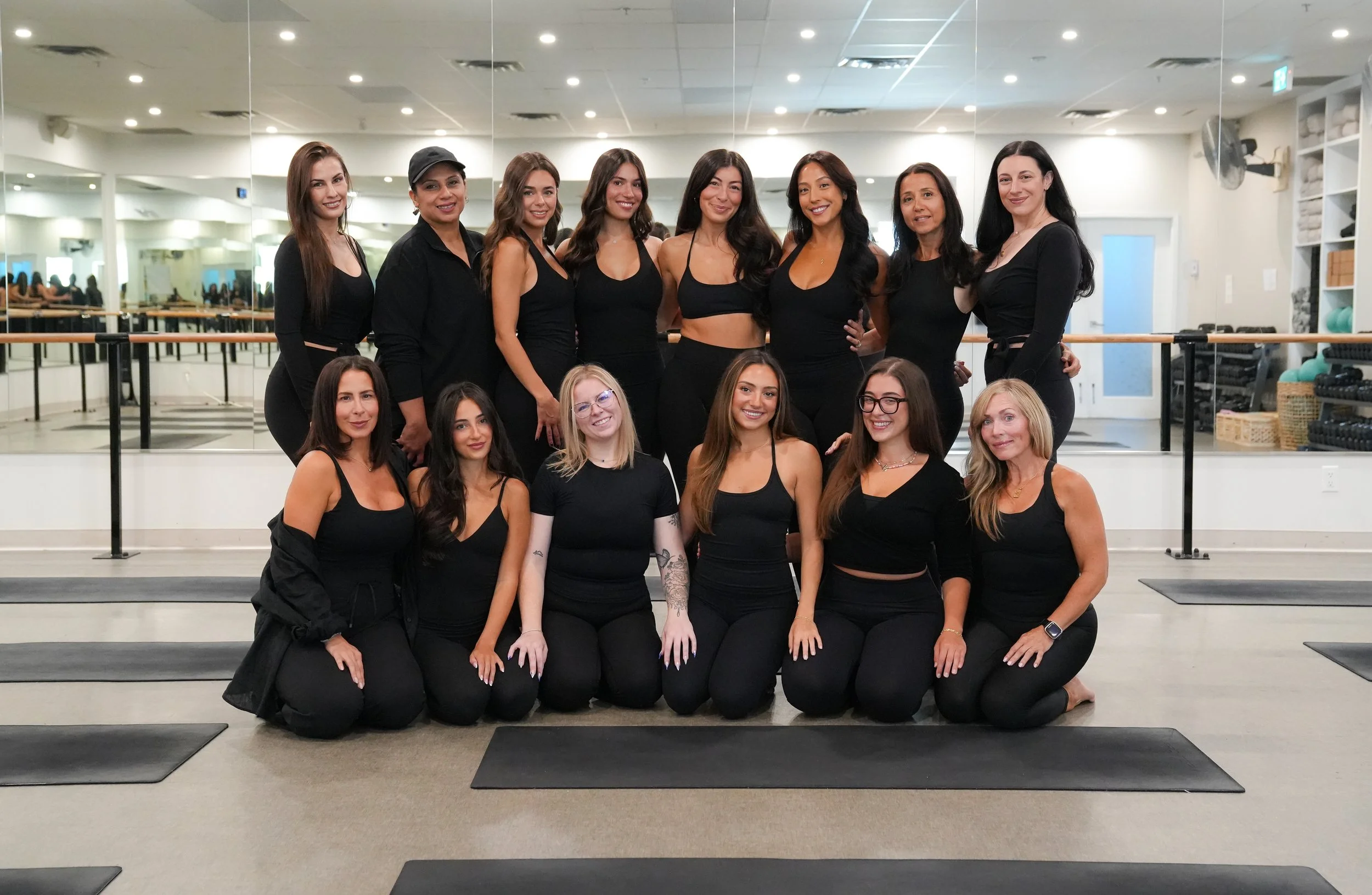 Group of women in workout clothes posing in a fitness studio with mirrors and exercise equipment in the background.