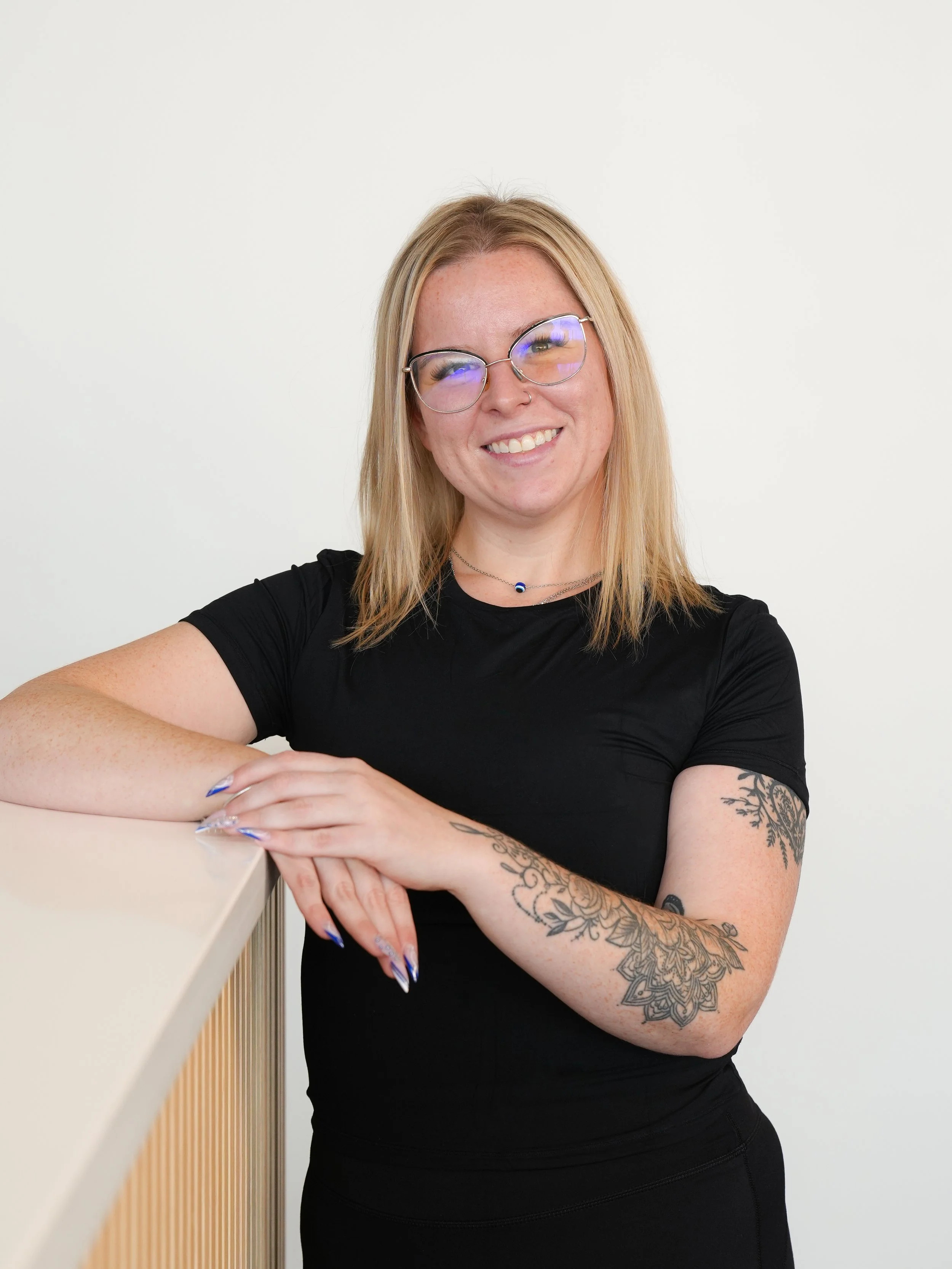 A young woman with blonde hair, glasses, and tattoos on her arms, smiling while leaning on a wooden railing against a plain white background.