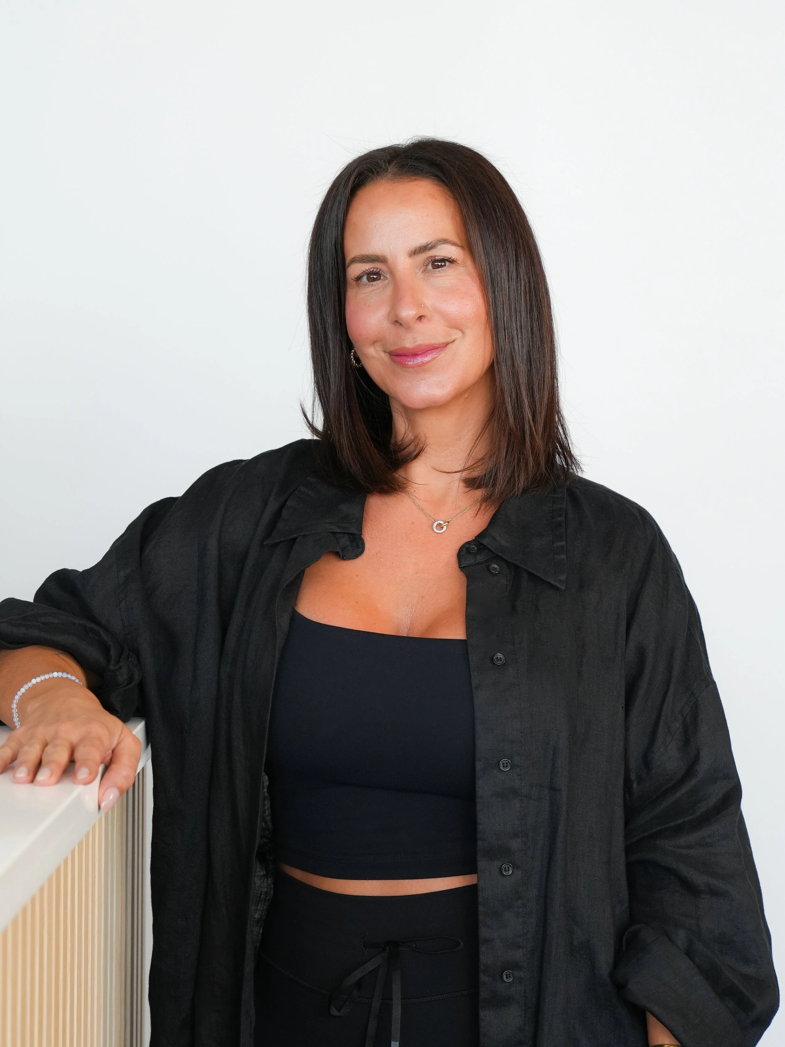 A woman with straight dark hair wearing a black shirt and a necklace, posing with a slight smile against a plain light background, resting her hand on a white surface.