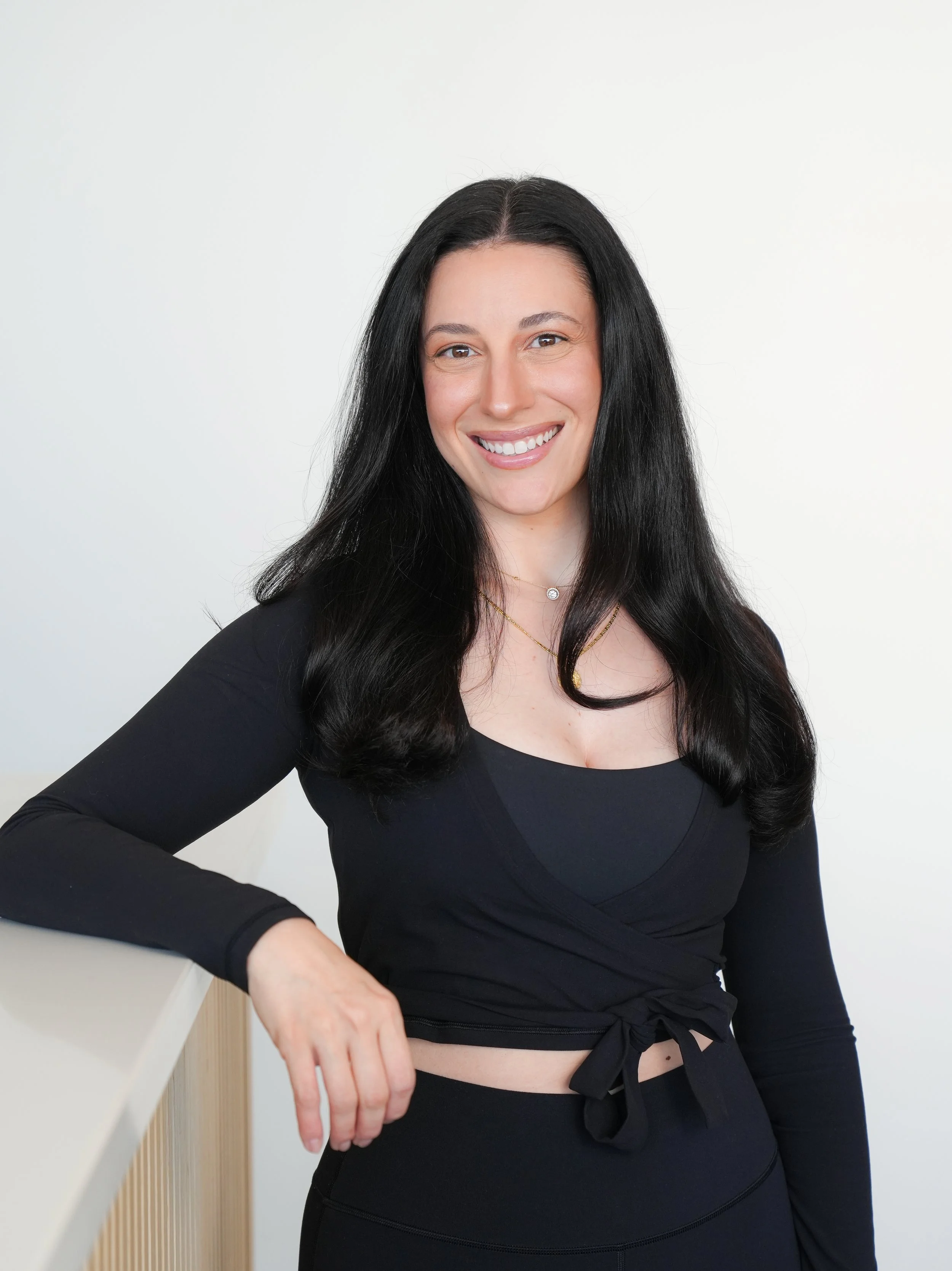 A woman with long black hair smiling, wearing a black top with a tied knot at the waist, standing against a plain white background.