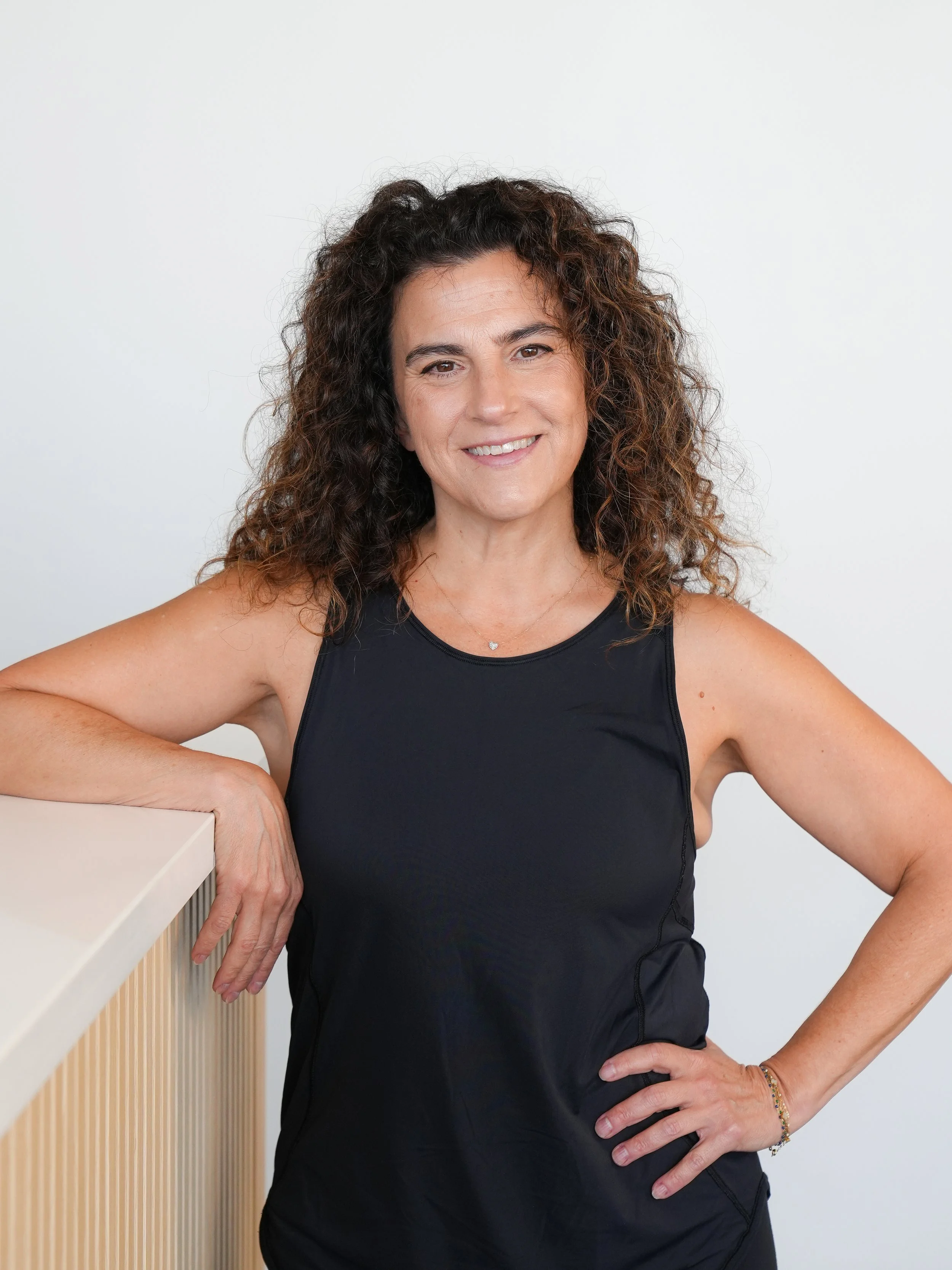 A woman with curly dark hair smiling, wearing a black sleeveless top, standing with one arm resting on a white surface, and the other hand on her hip, against a white background.