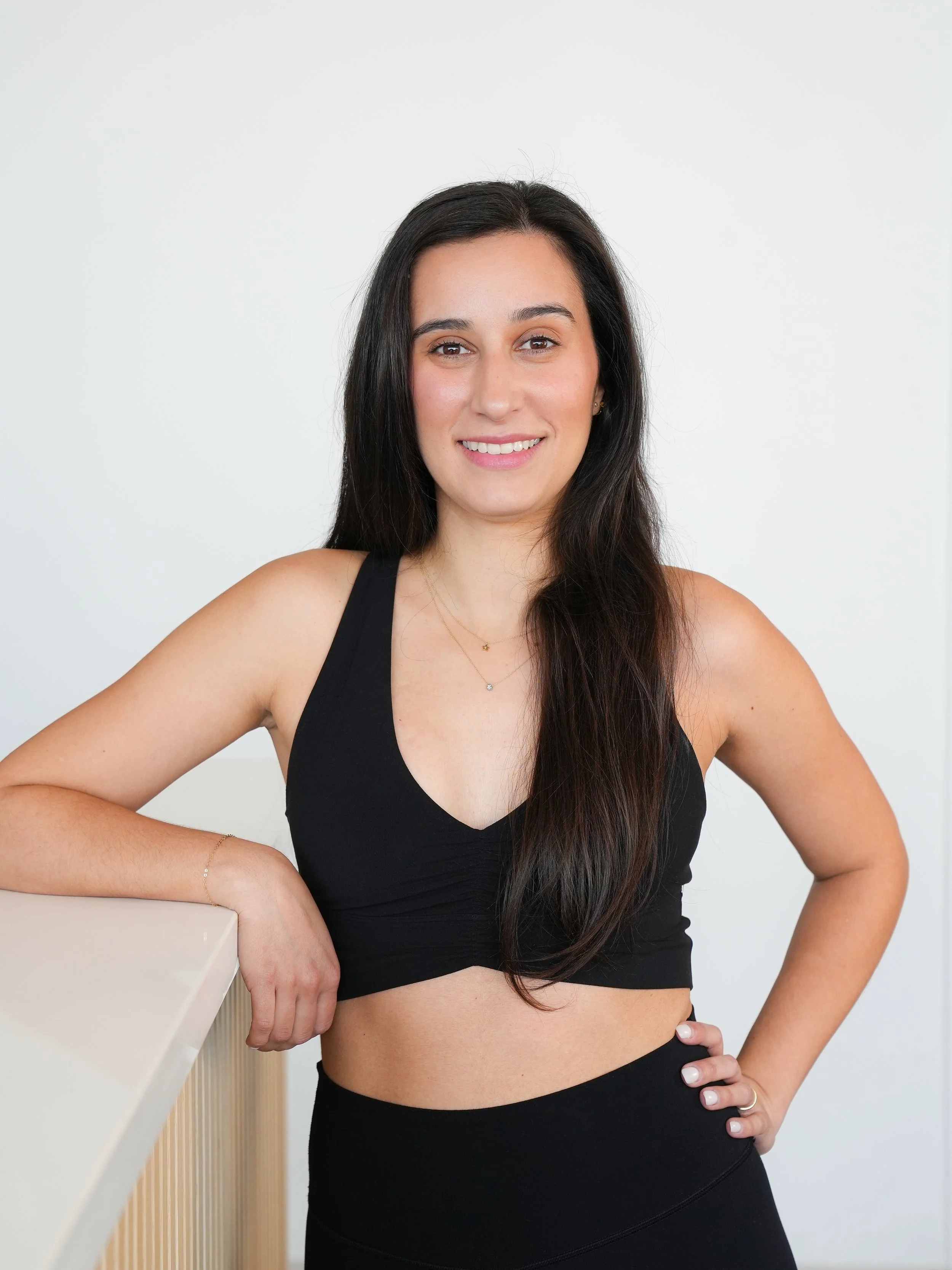 A young woman with long dark hair wearing a black sports bra and high-waisted leggings, leaning on a white surface with her left arm, smiling at the camera, with a plain white wall background.