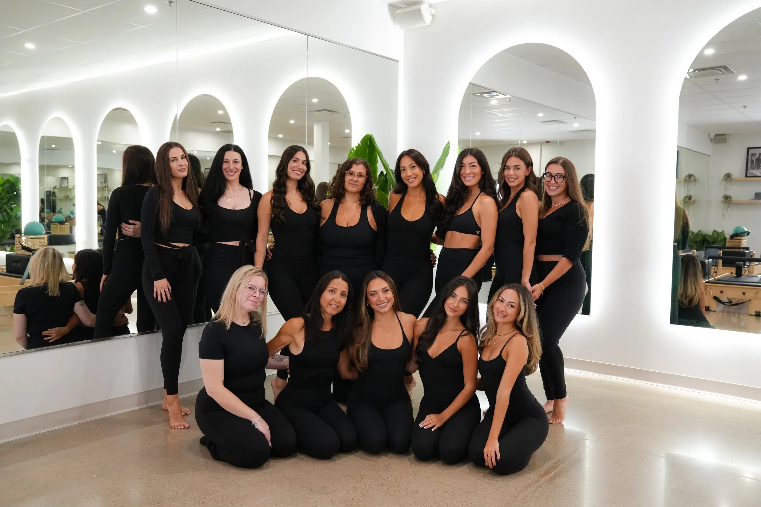 Group of women dressed in black posing in front of a large mirror in a modern indoor space with white arches and plants.