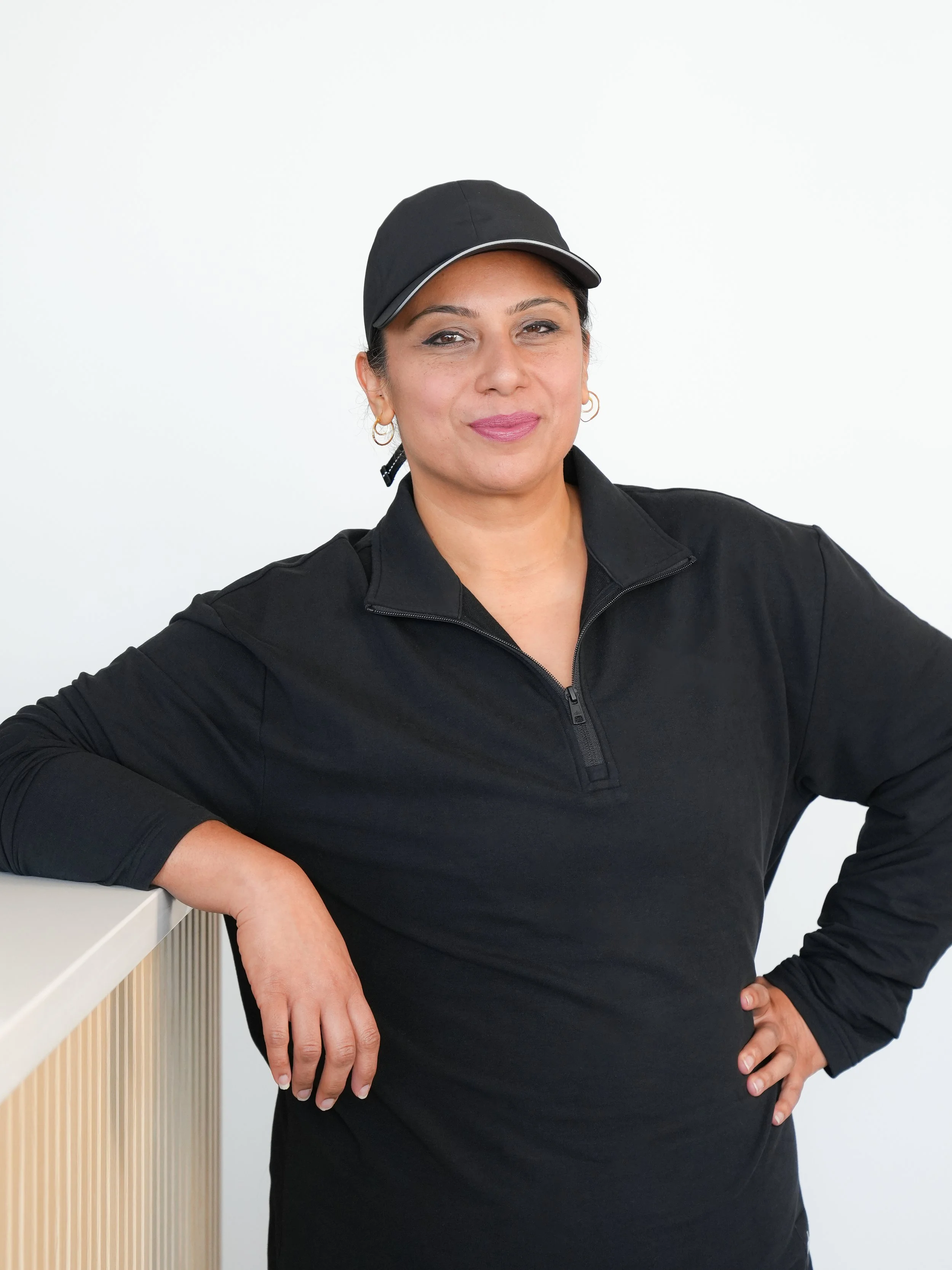 A woman in a black athletic jacket and cap leaning on a white surface against a plain white background.