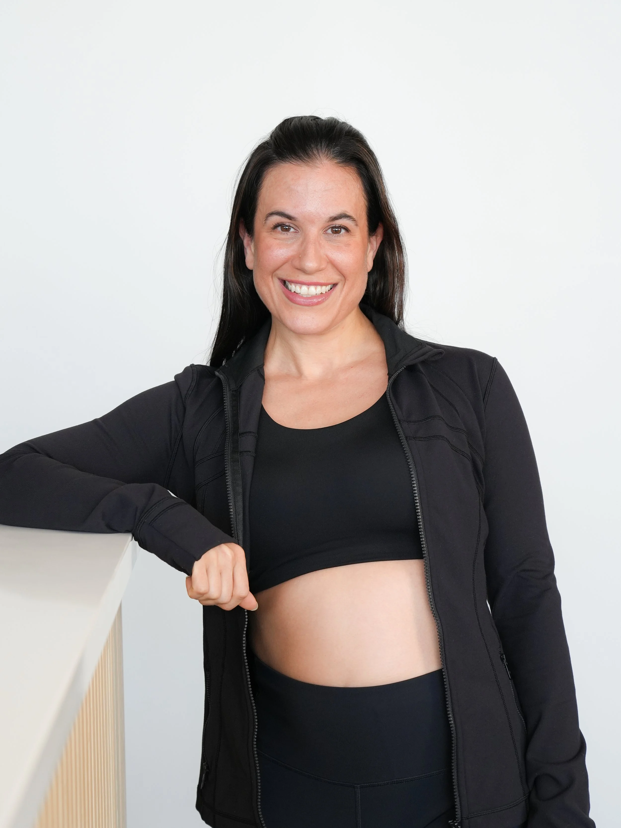 A woman with long dark hair smiling, wearing a black athletic jacket and black workout top, standing against a plain white background, with her left arm resting on a white counter.
