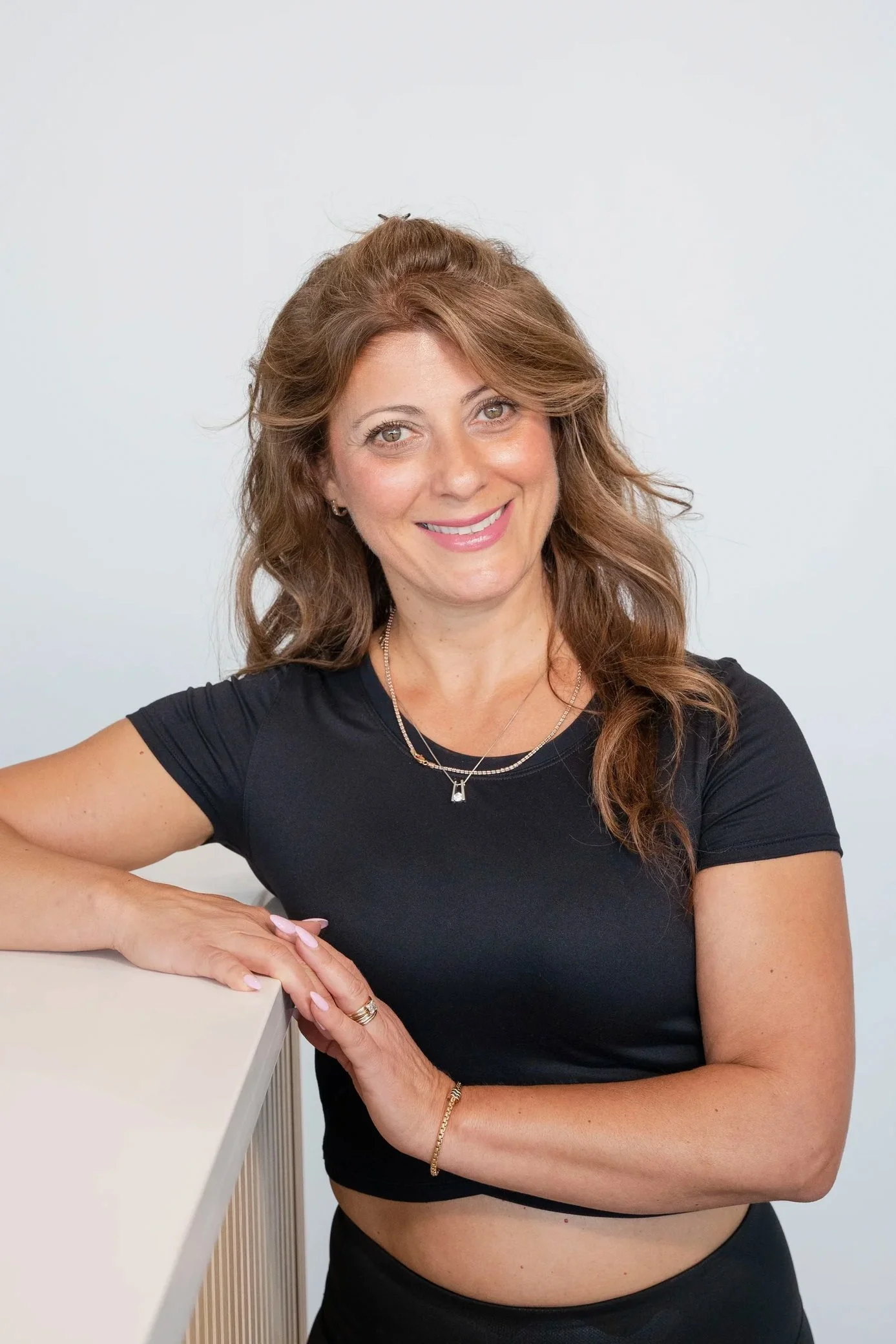 A smiling woman with wavy brown hair, wearing a black crop top and jewelry, resting her arm on a white surface, against a plain light background.