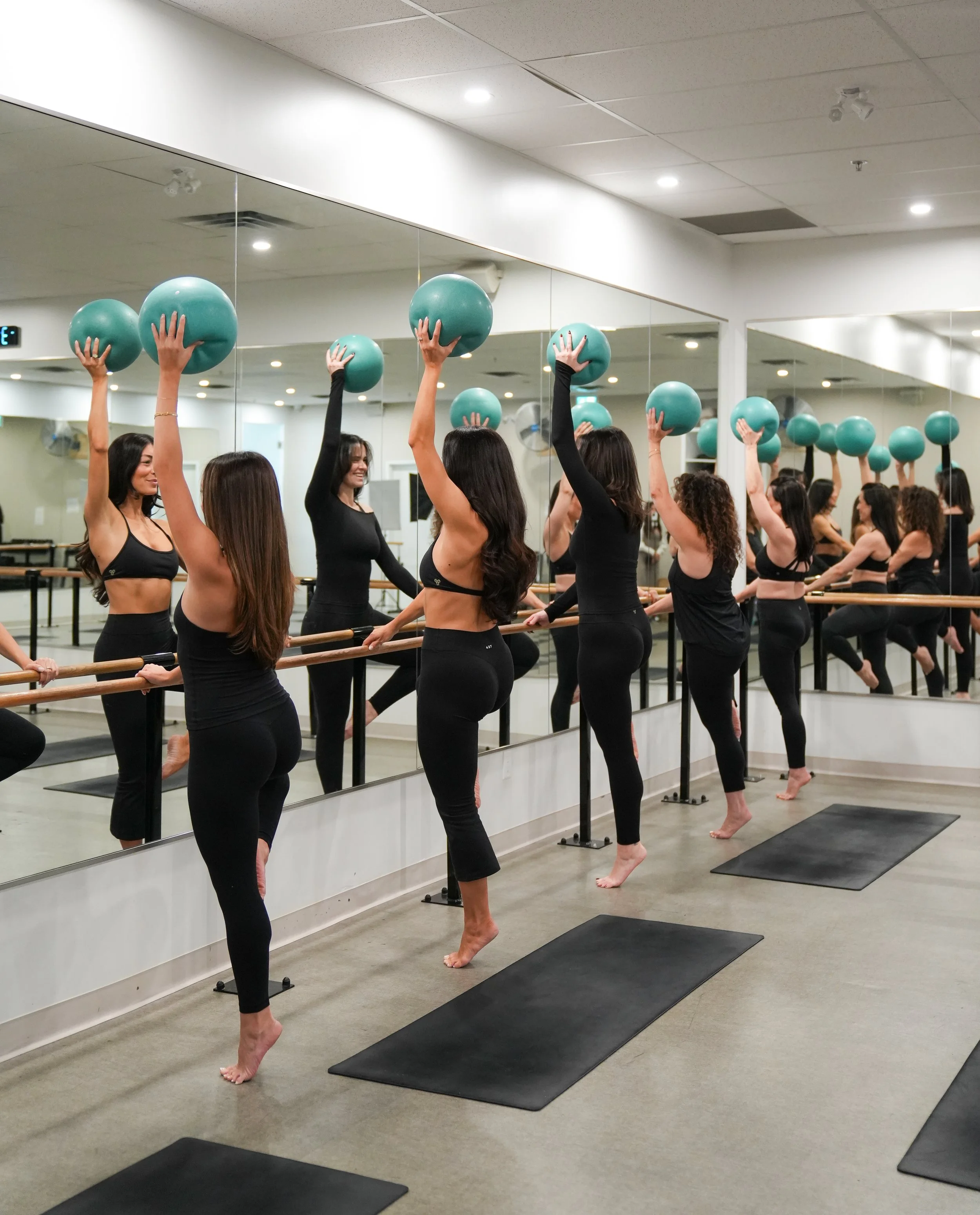 Women participating in a fitness class in a dance studio, holding teal exercise balls, standing on black mats, using ballet barres, with mirrors on the wall, and a ceiling with multiple lights and vents.