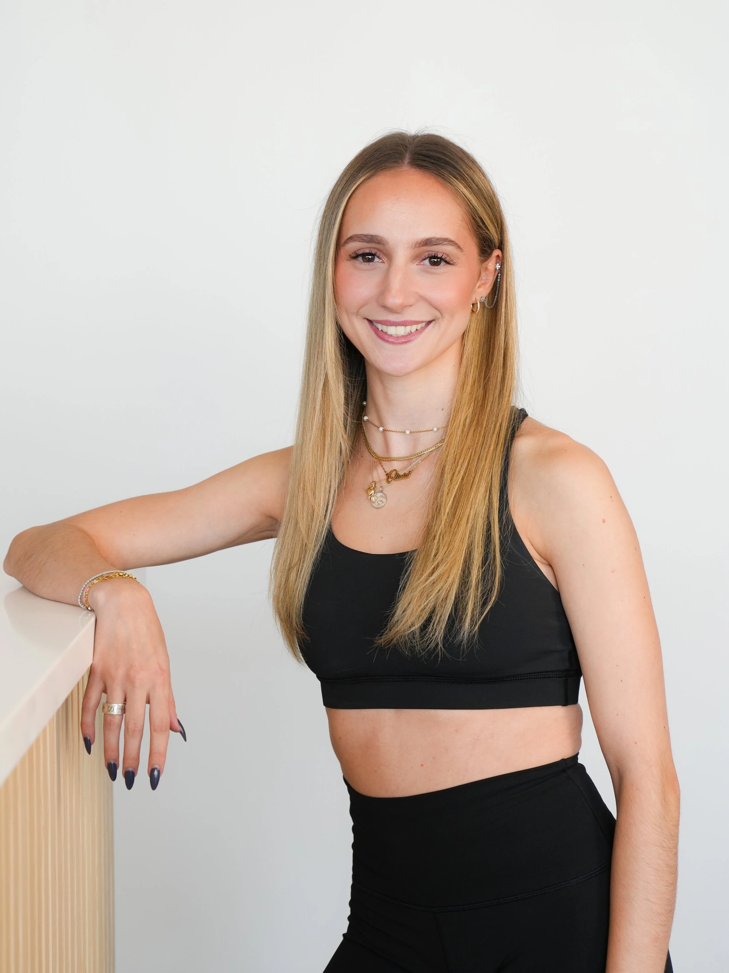 A young woman with long blonde hair wearing a black sports bra and matching pants, accessorized with jewelry, smiling, with her right arm resting on a wooden surface.