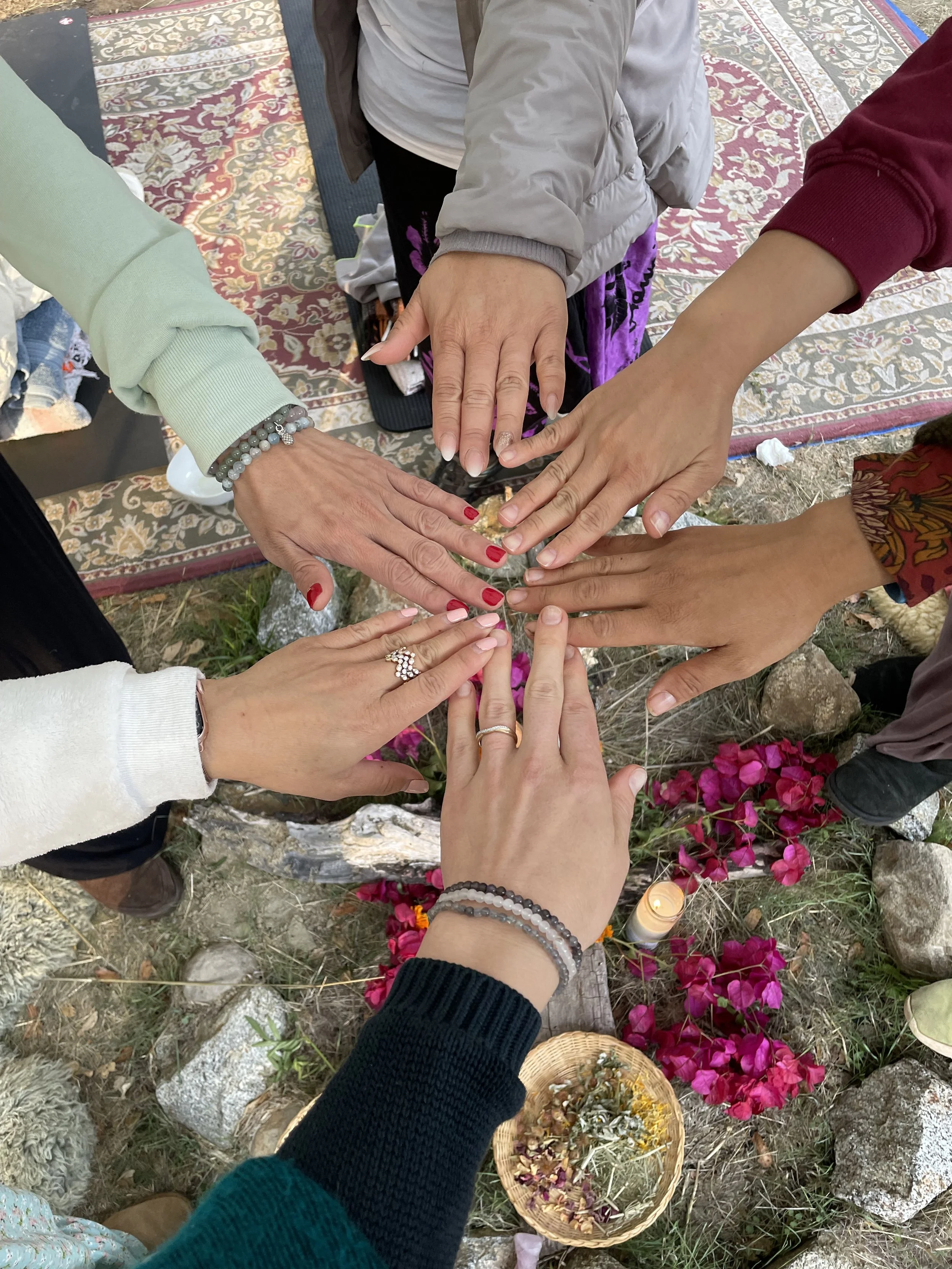 Five people with their hands stacked together in a circle, surrounded by flowers, rocks, and a lit candle outdoors.