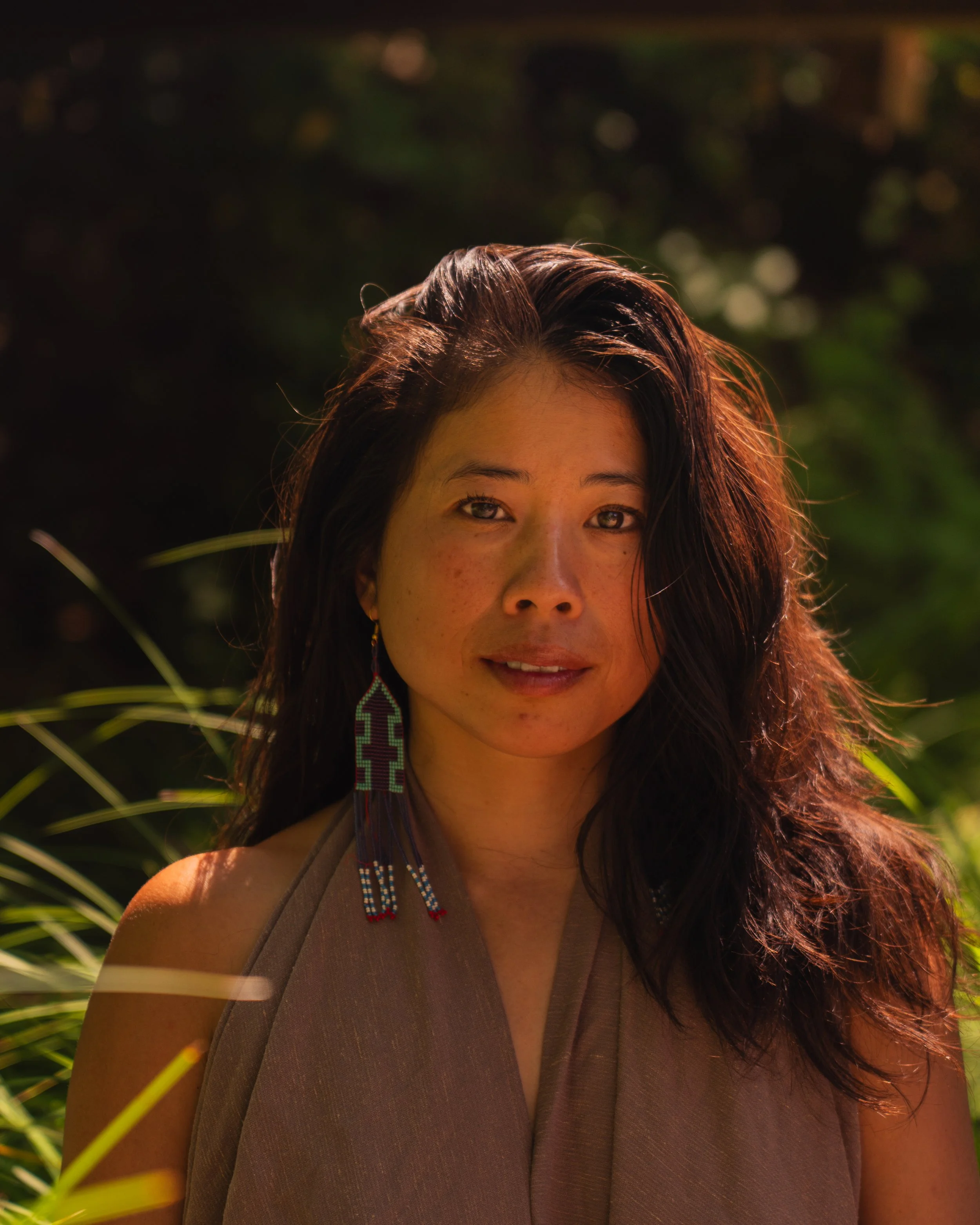 Portrait of a woman with long dark hair, wearing a brown top and beaded earring, outdoors in natural light.