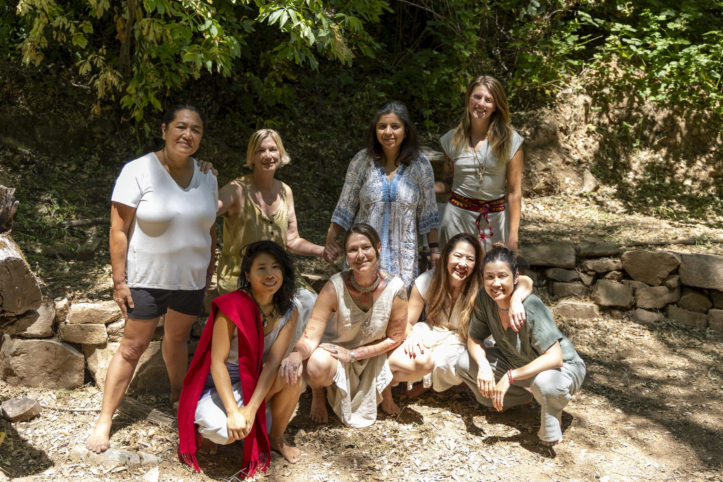 Group of nine women standing and sitting outdoors in a wooded area, smiling and posing for a photo.