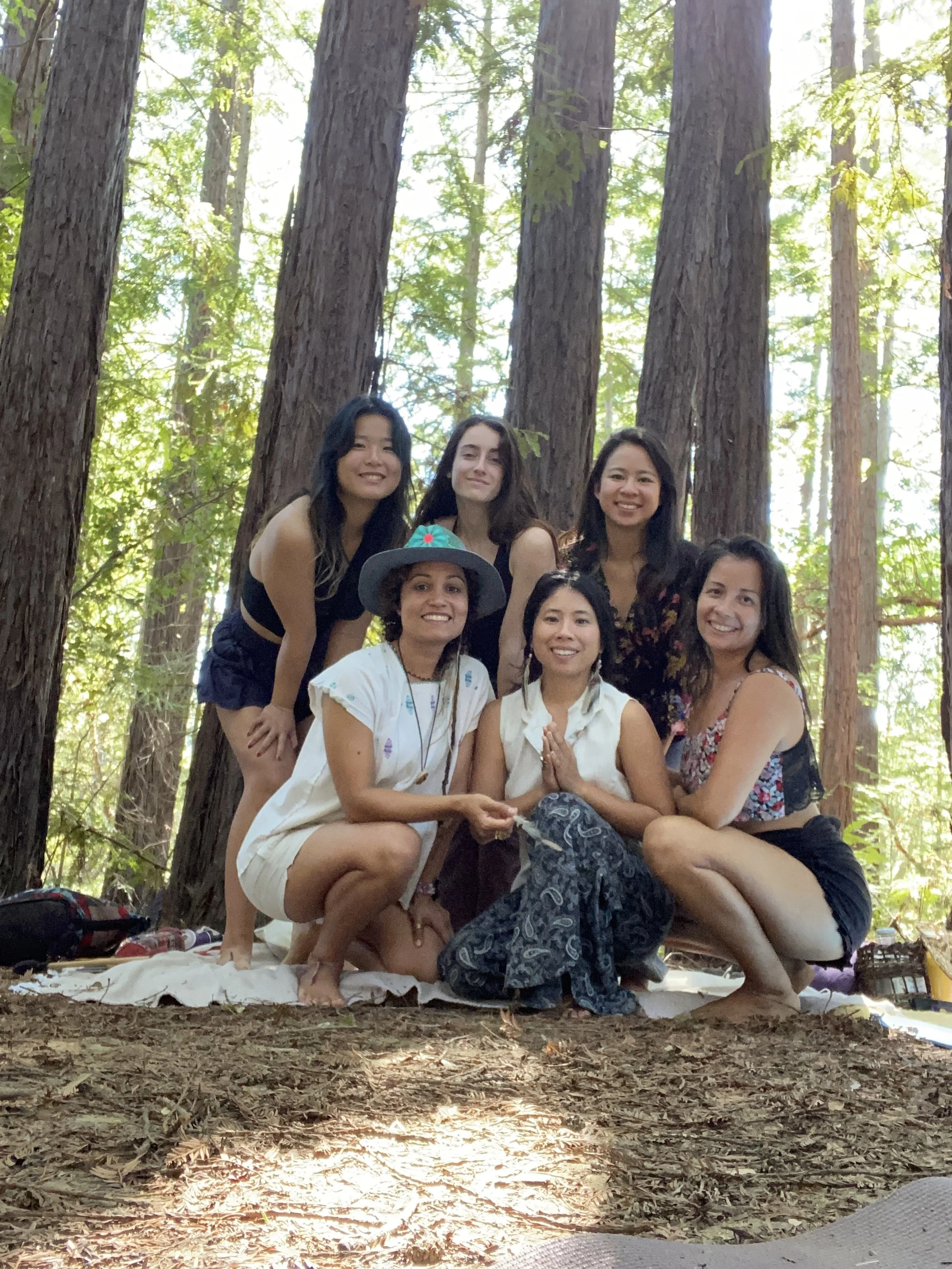 Seven women sitting and kneeling on a blanket in a forest with tall trees, smiling and posing for the photo.