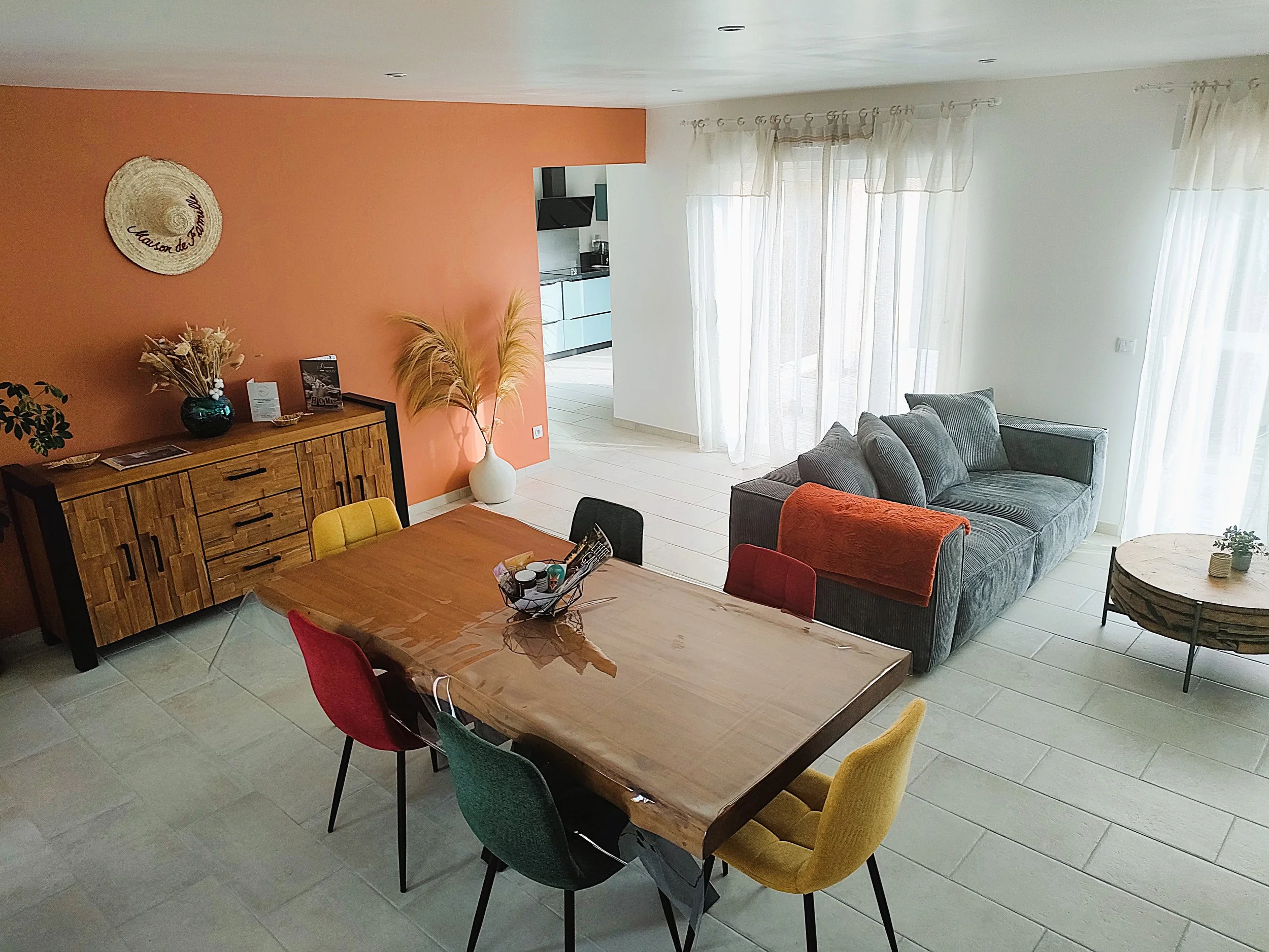 Salon lumineux avec un canapé gris, une table en bois avec des chaises colorées, et une grande fenêtre avec des rideaux blancs, cuisine en arrière-plan.