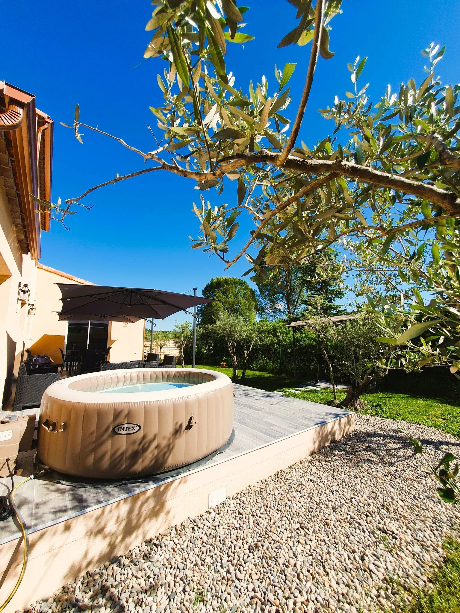 Piscine gonflable dans un jardin avec arbres, terrasse, parasol, et un ciel bleu clair.
