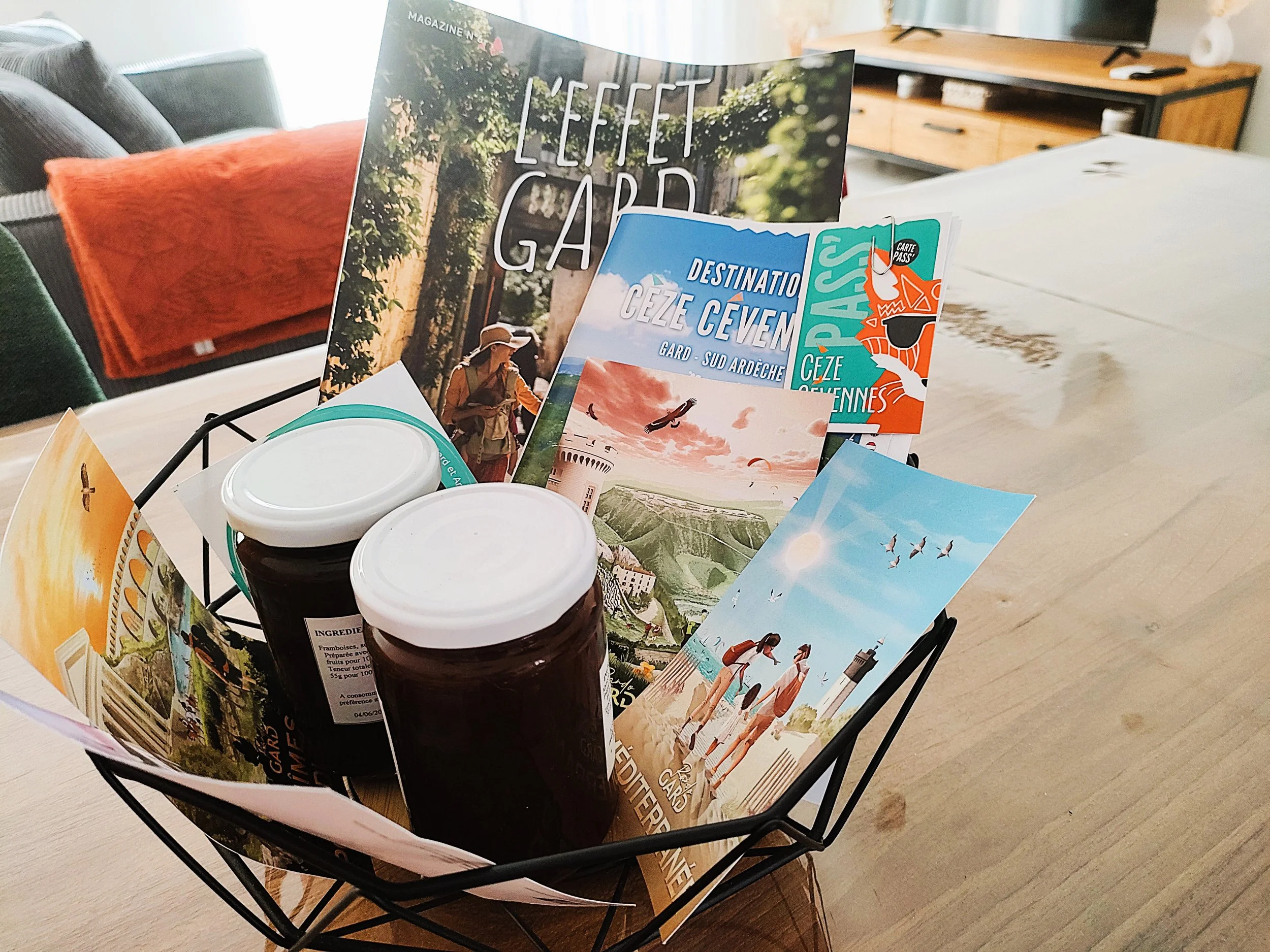 Panier contenant des dépliants touristiques de la région de Garde-Sud-Ardèche, deux pots de confiture, posé sur une table en bois dans un intérieur moderne.
